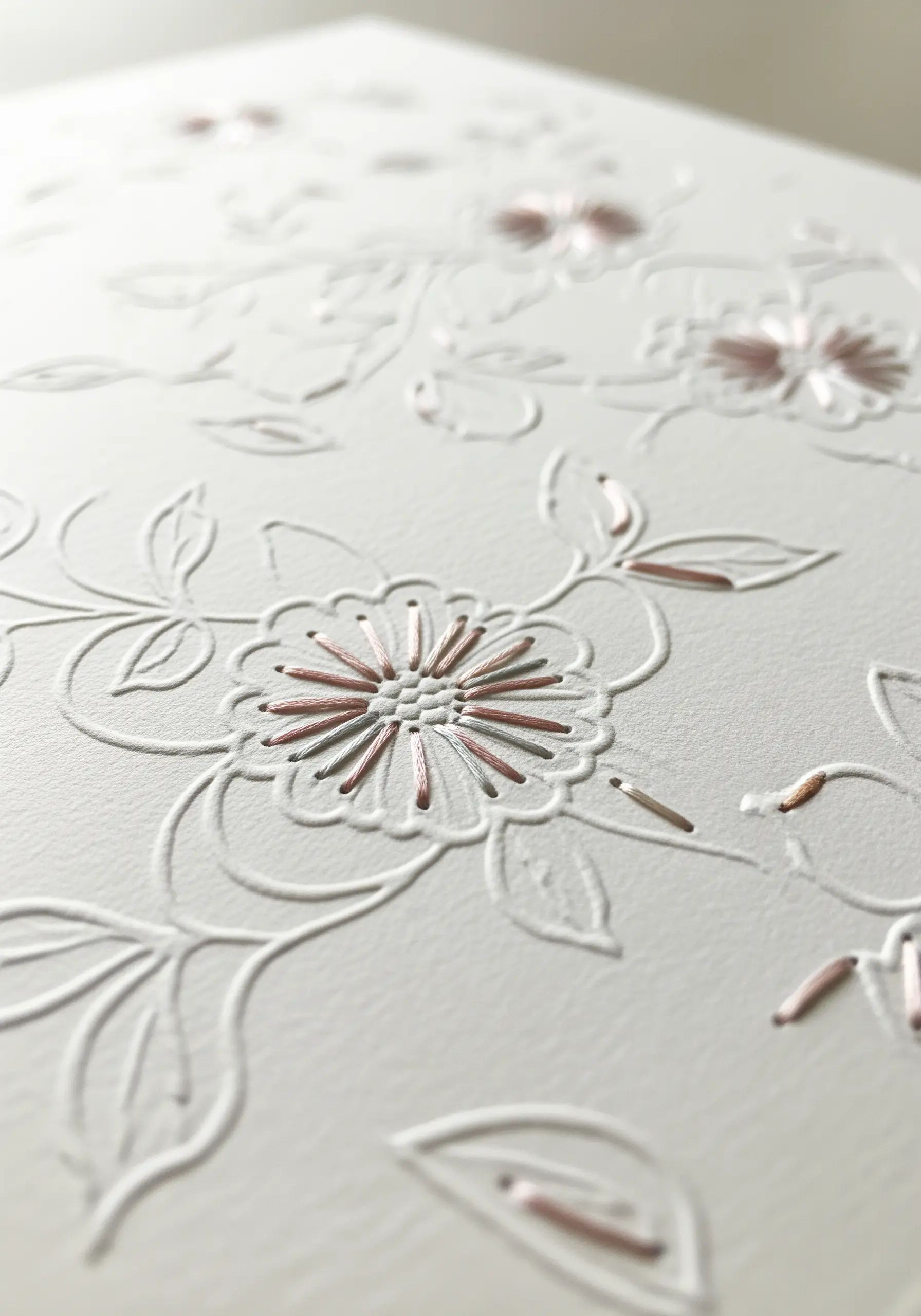 A close-up of a white floral embossed paper design with small, selective details stitched in pink thread.
