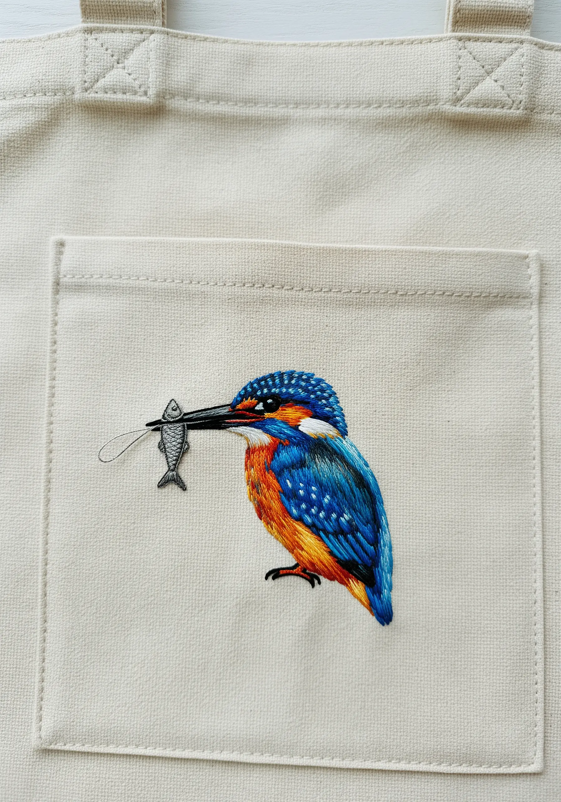 Detailed embroidery of a kingfisher with a fish on the pocket of a canvas tote bag.