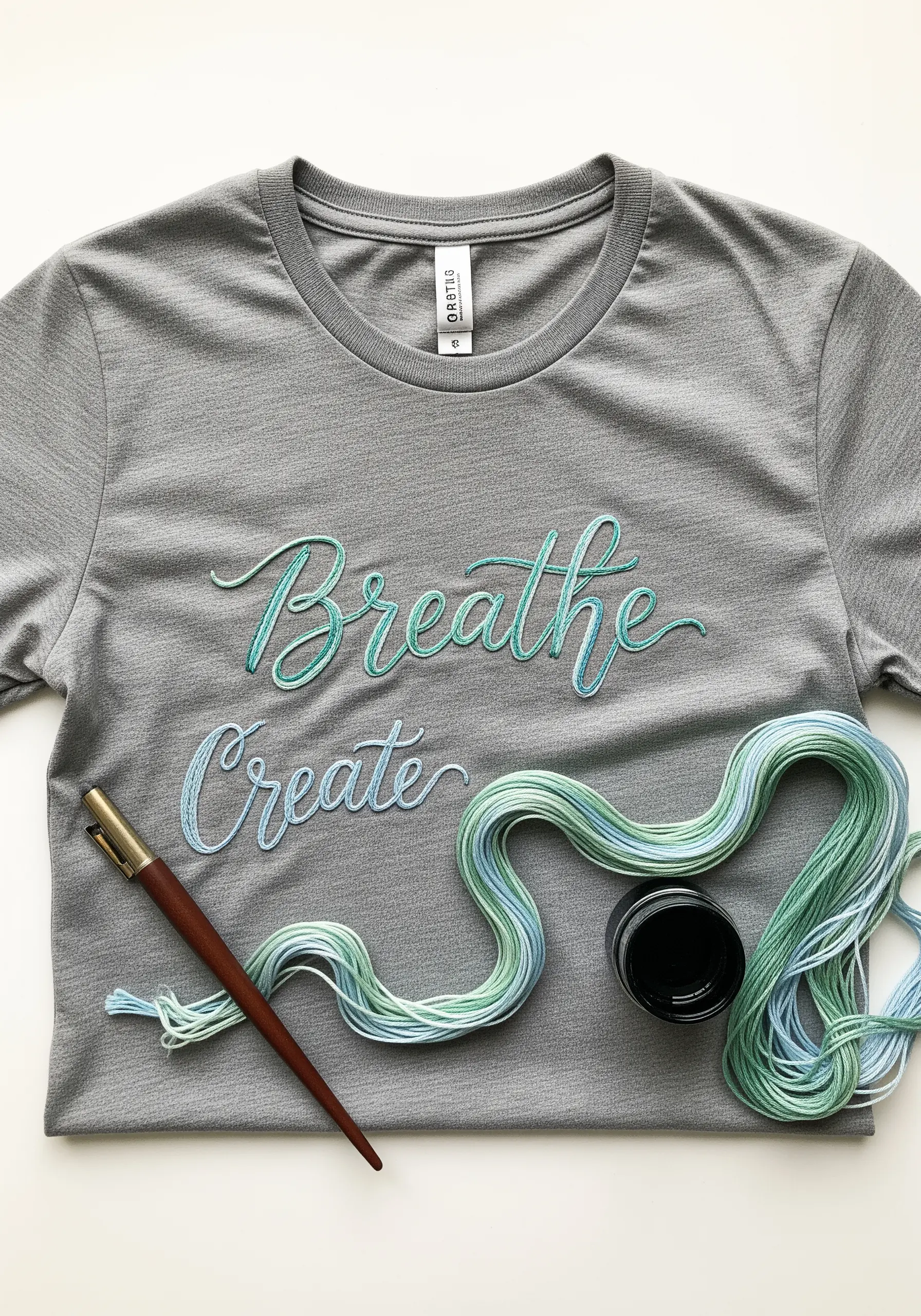 The words 'Breathe Create' embroidered in an ombré blue-green variegated thread on a gray t-shirt.