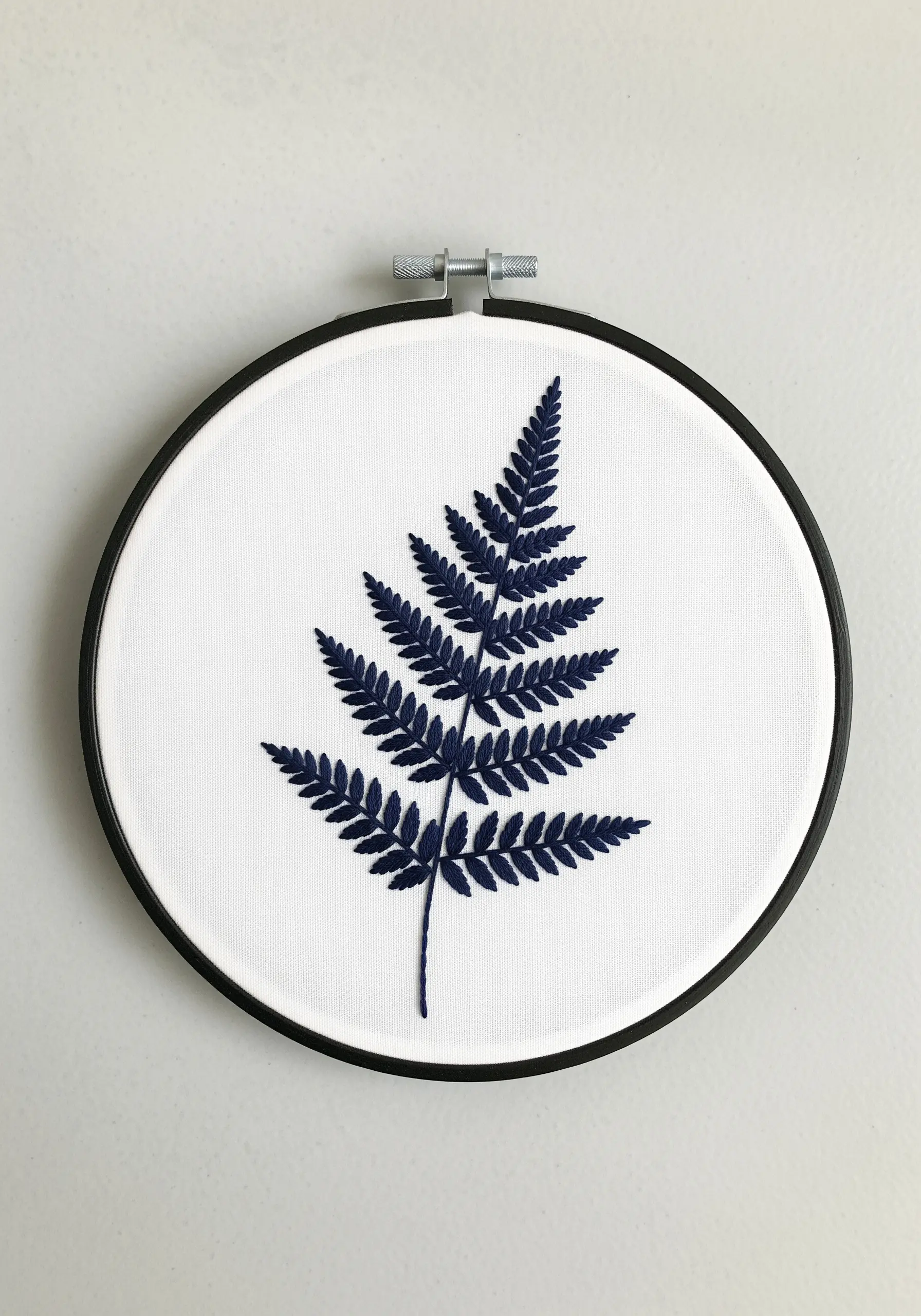 Detailed navy blue embroidery of a fern leaf on white fabric in a black hoop.