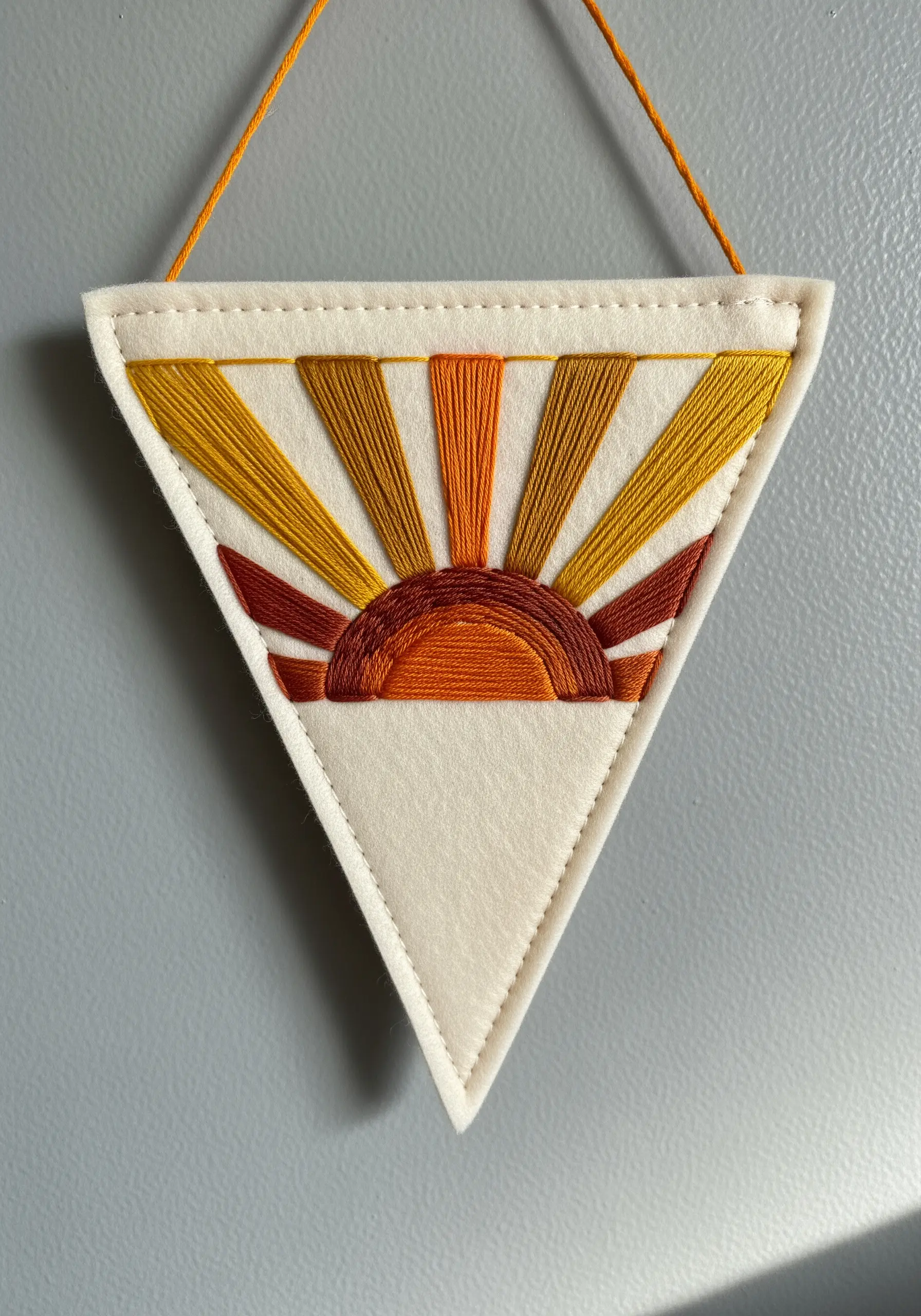 Satin-stitched sunburst design in warm tones on a cream felt pennant.