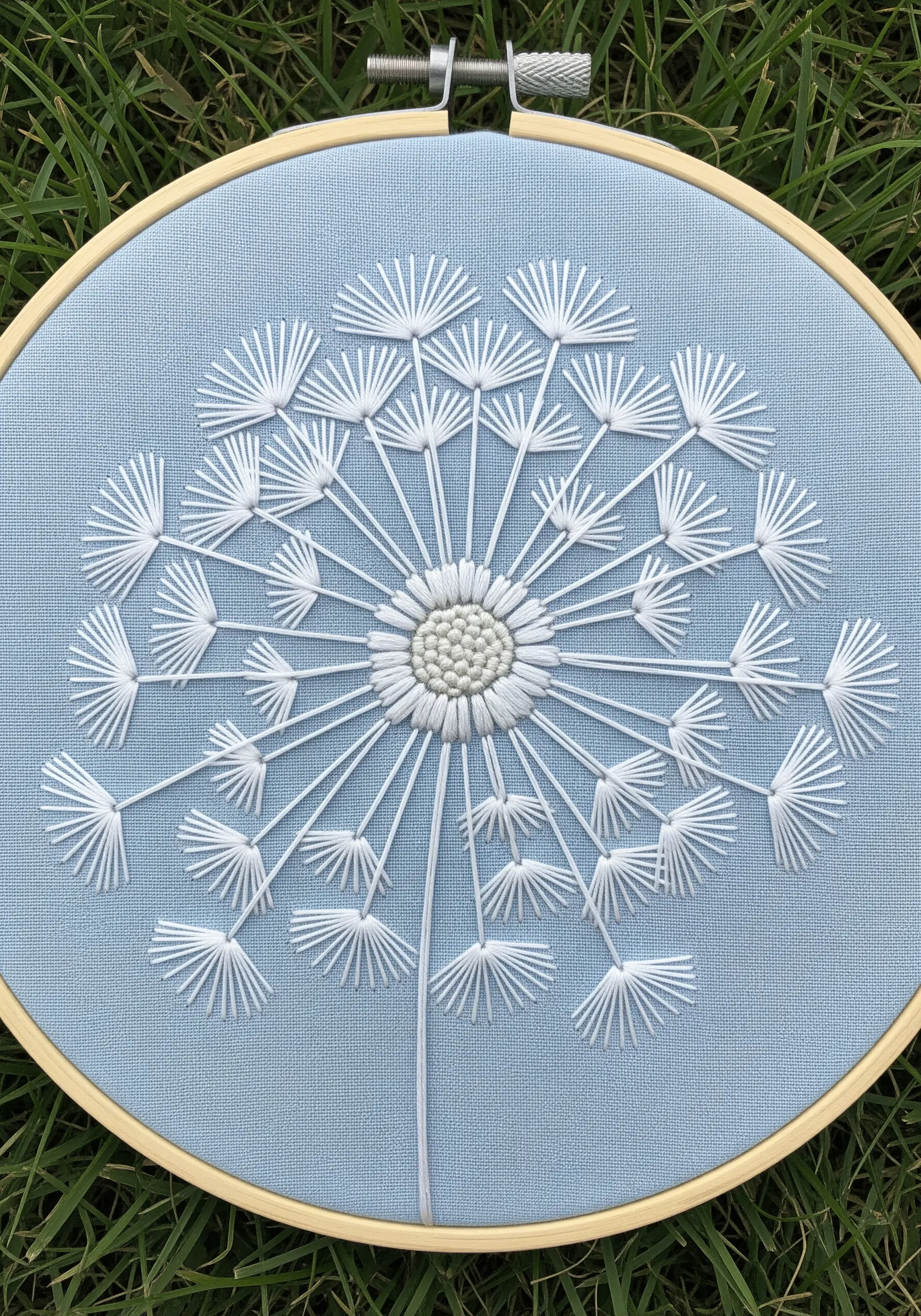 A delicate dandelion head embroidered in white thread on light blue fabric.