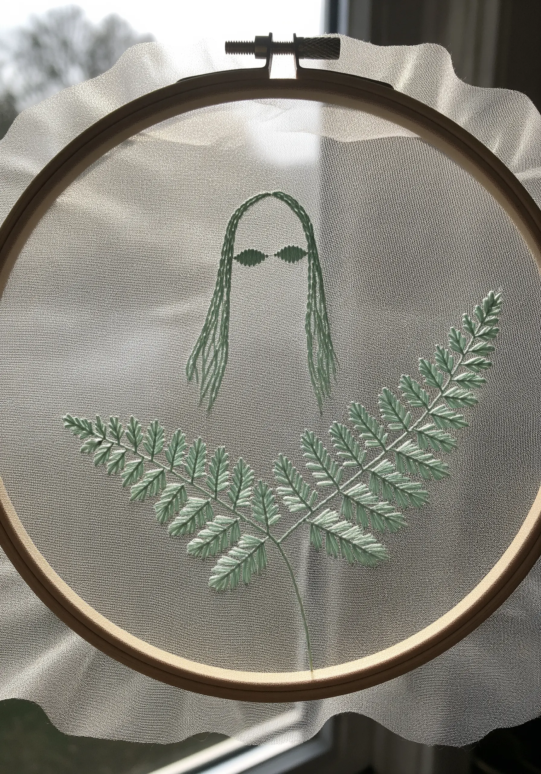 A delicate mint green fern embroidered on translucent tulle fabric in a hoop.
