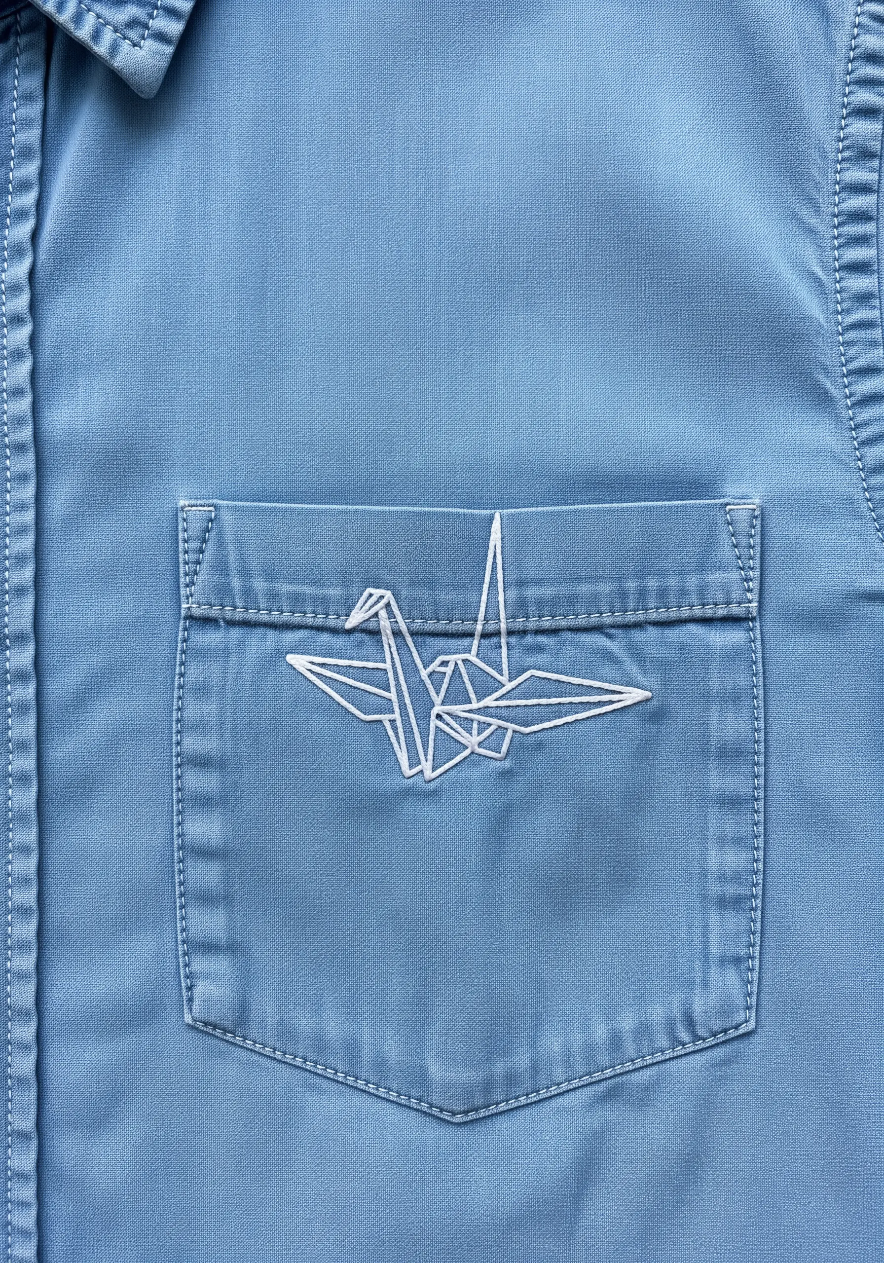 A geometric origami crane embroidered in white thread on the pocket of a blue chambray shirt.