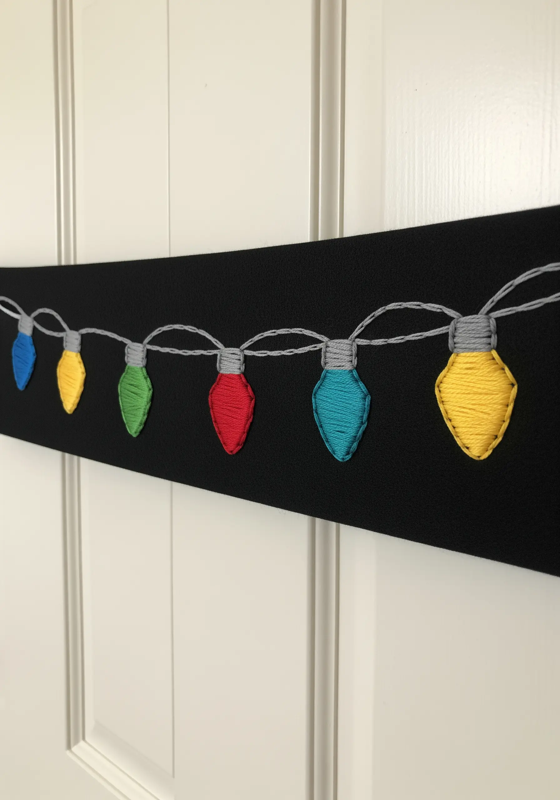 A string of colorful embroidered Christmas lightbulbs on black fabric.