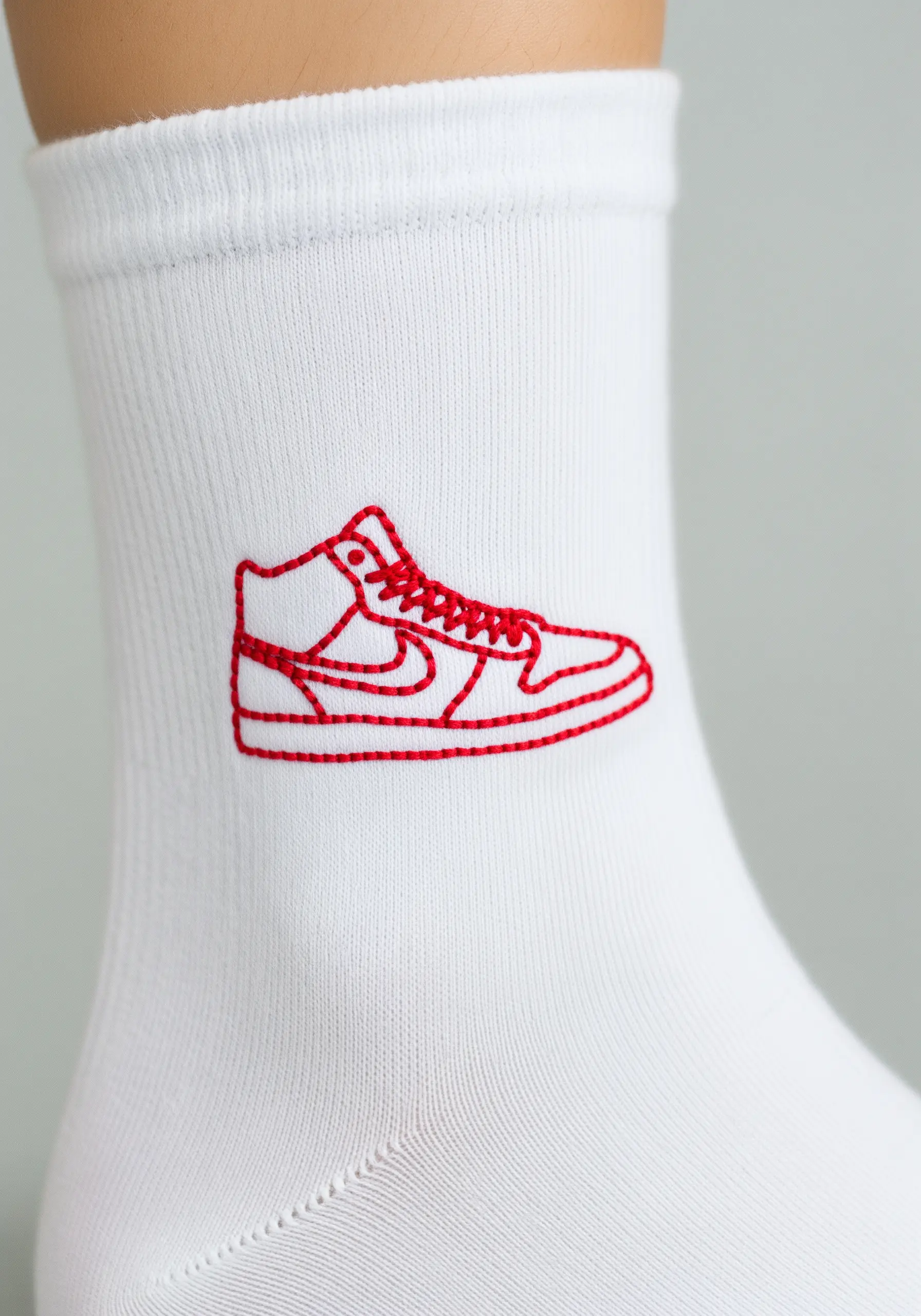 A red line-art sneaker embroidered on the side of a white knit sock.
