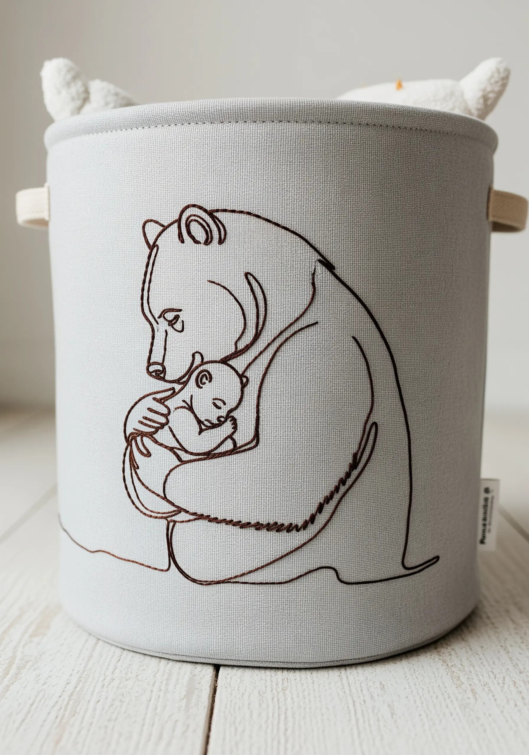 Line art of a mother bear hugging a cub, embroidered on the side of a grey fabric storage bin.