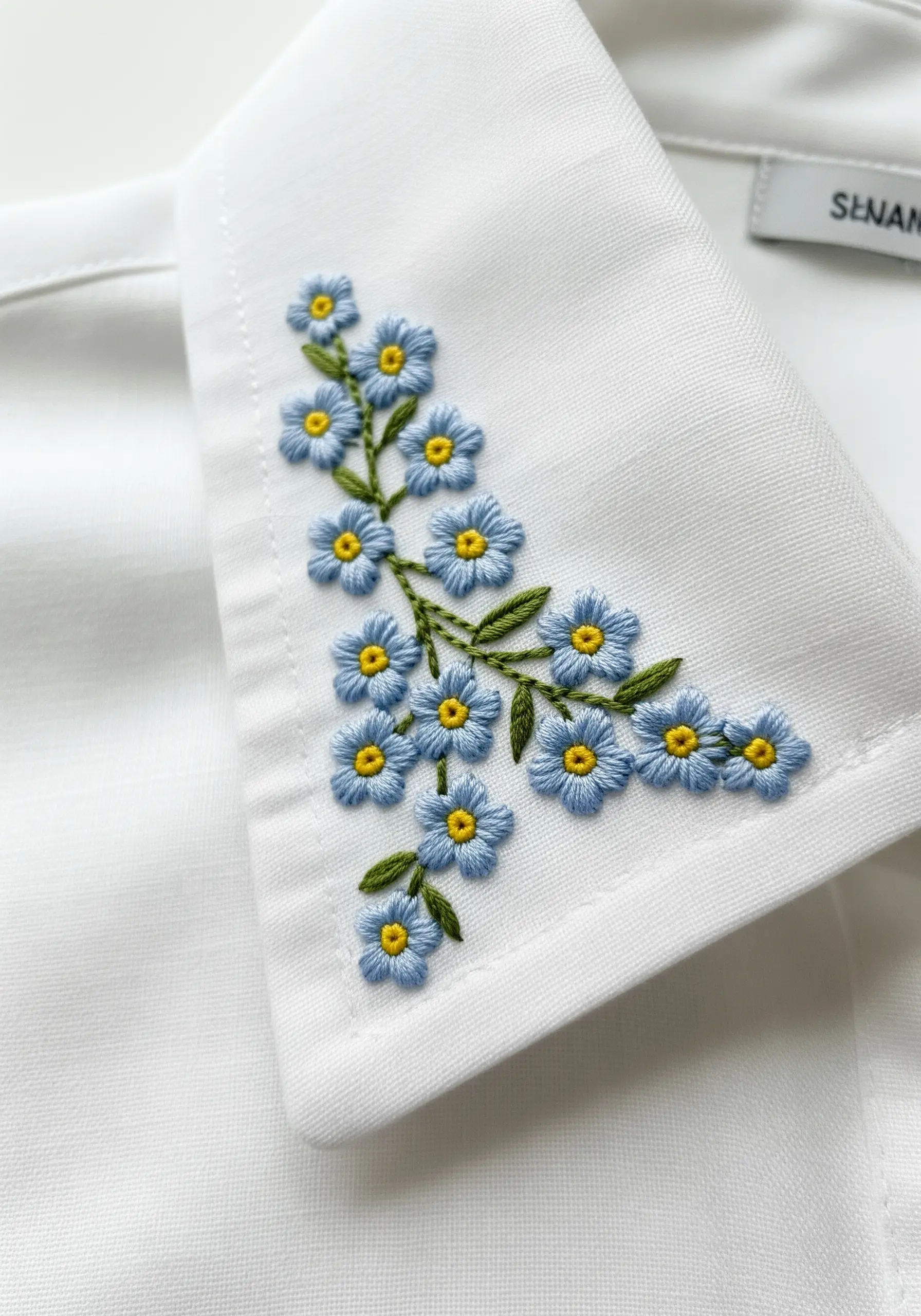 Delicate blue forget-me-not flowers with yellow centers embroidered on a white shirt collar.