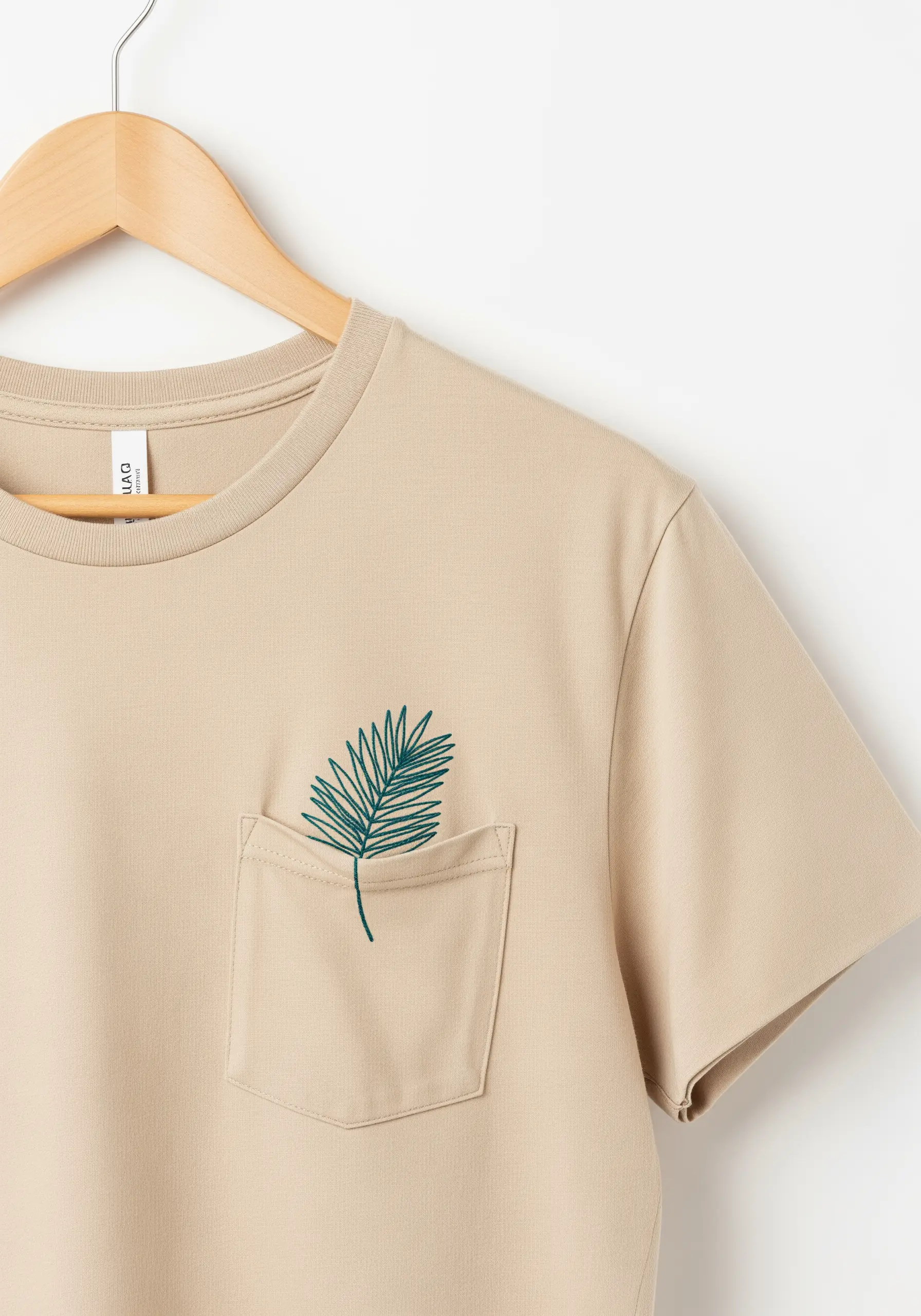 A single palm frond in teal thread embroidered on the pocket of a beige t-shirt.