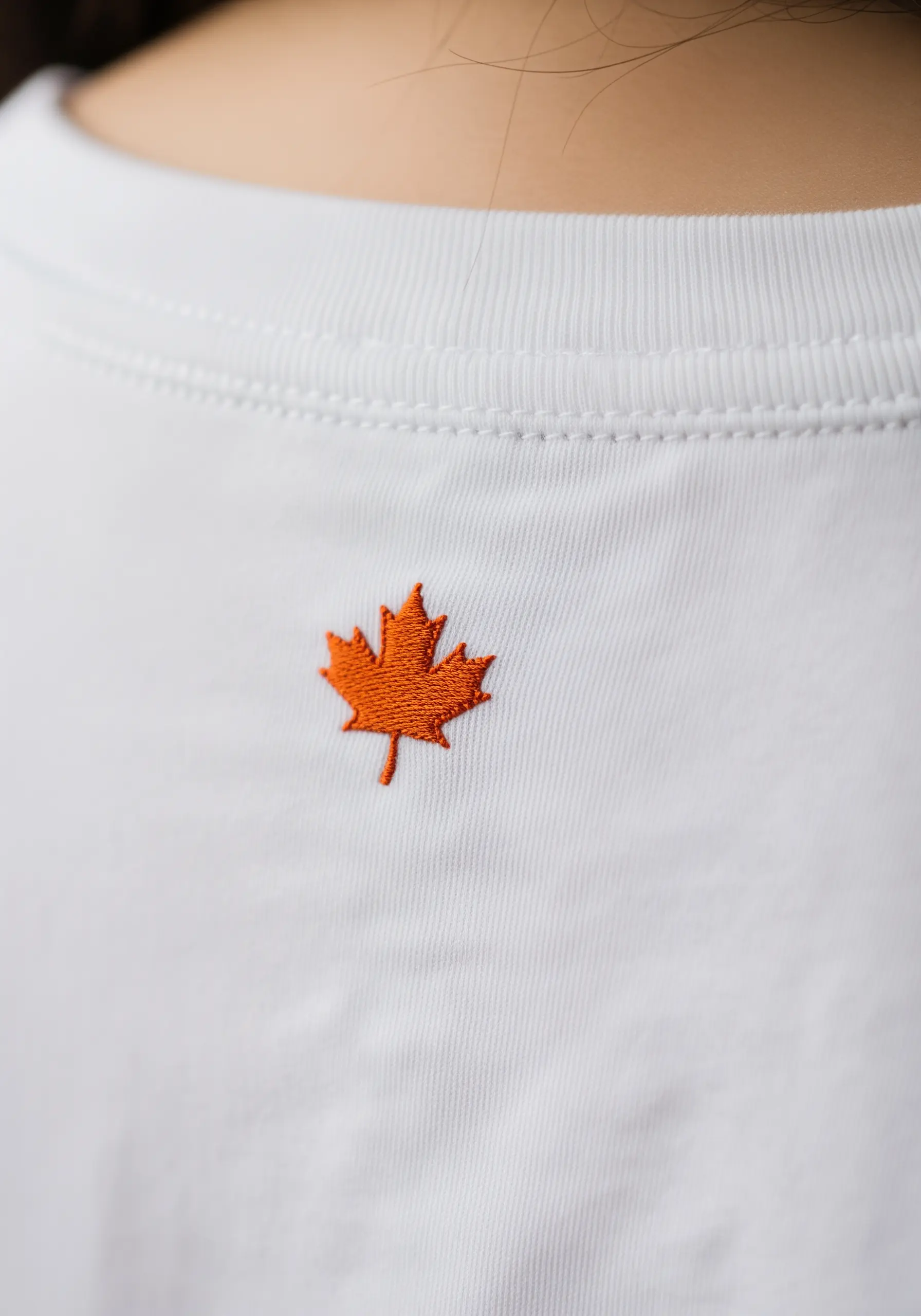 Small orange maple leaf embroidered on the back of a t-shirt, below the collar.