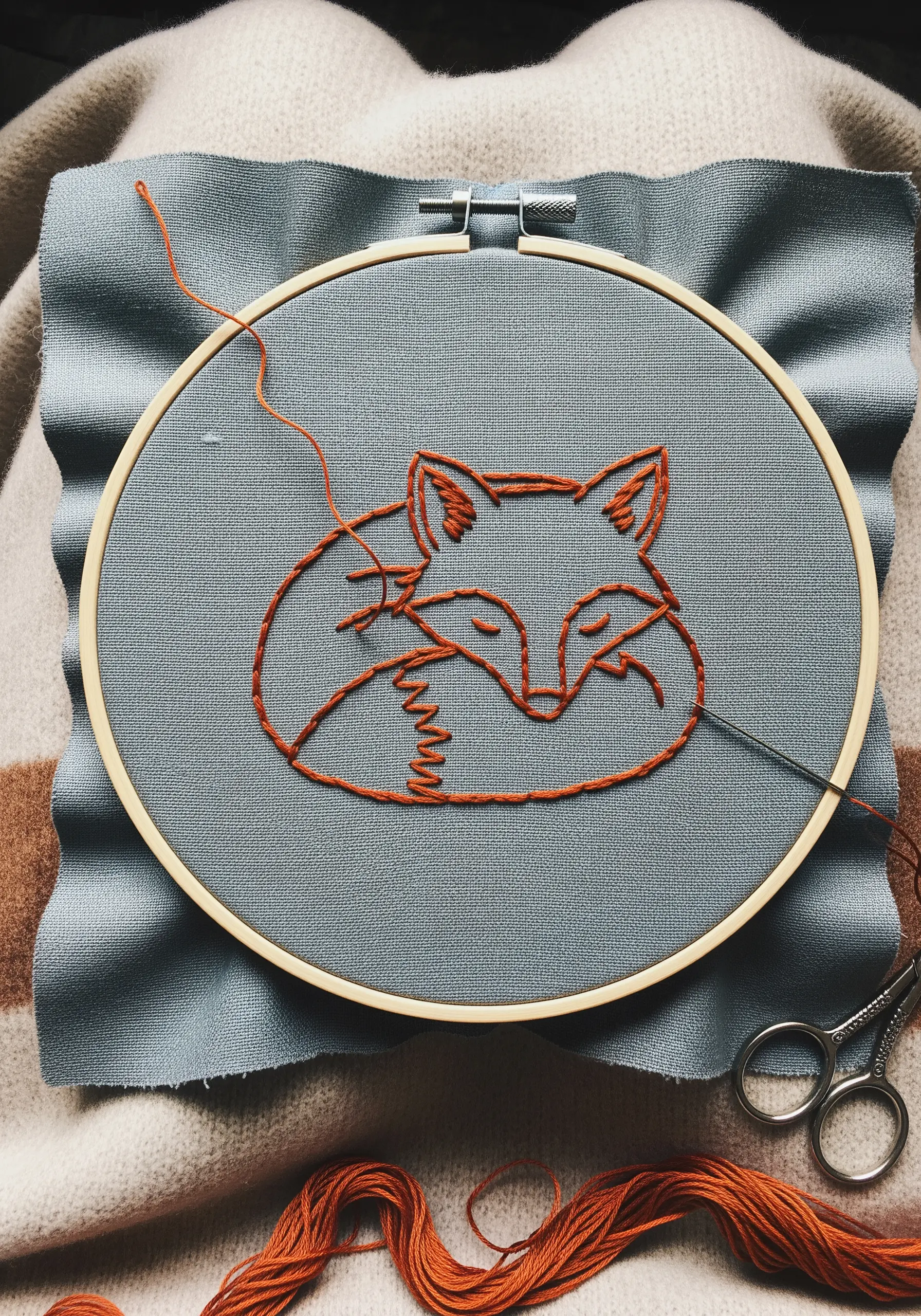 A line art sleeping fox in orange thread on blue fabric, showing varied stitch textures.