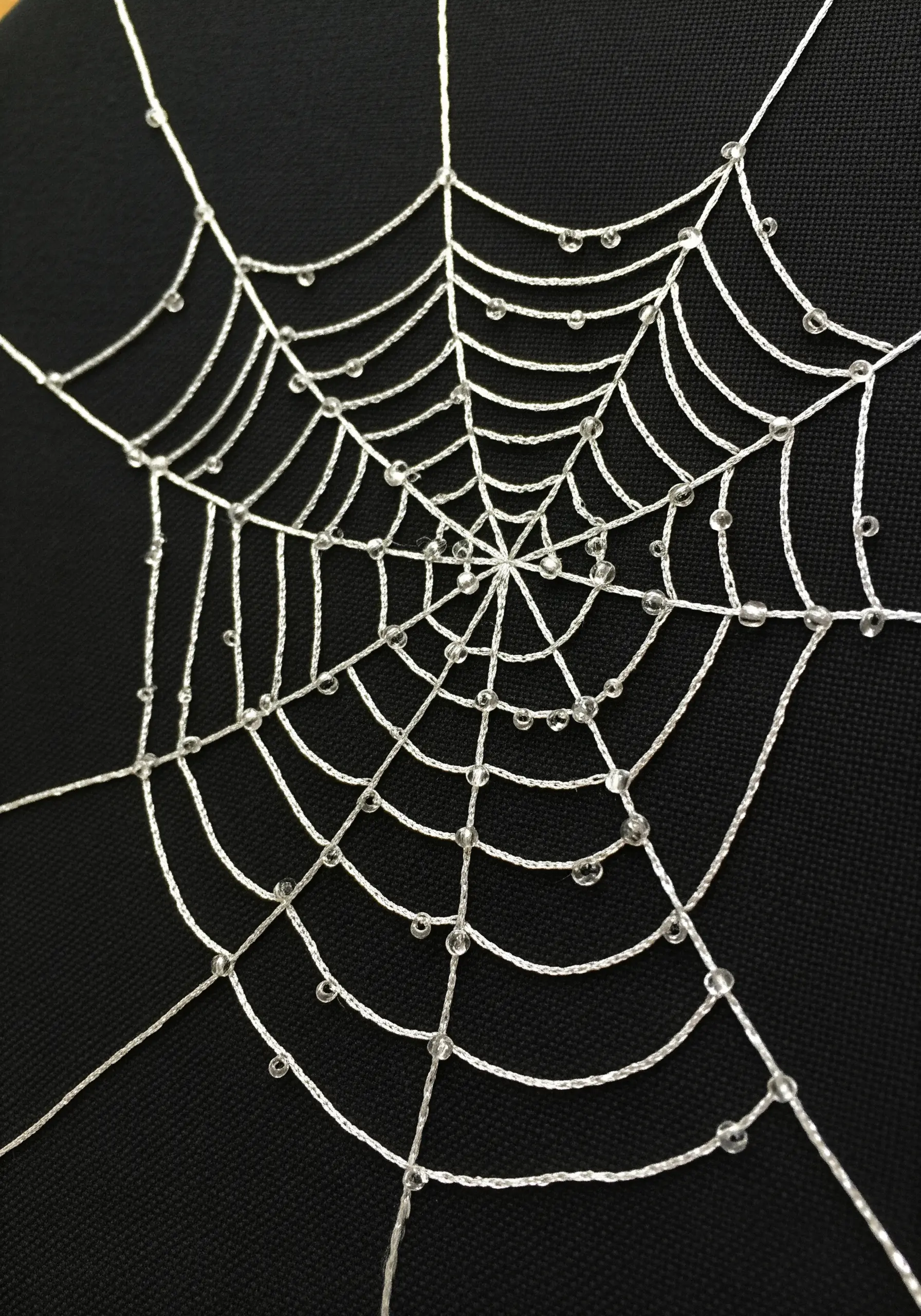 A delicate spiderweb embroidered in white on black fabric, adorned with tiny clear beads as dewdrops.