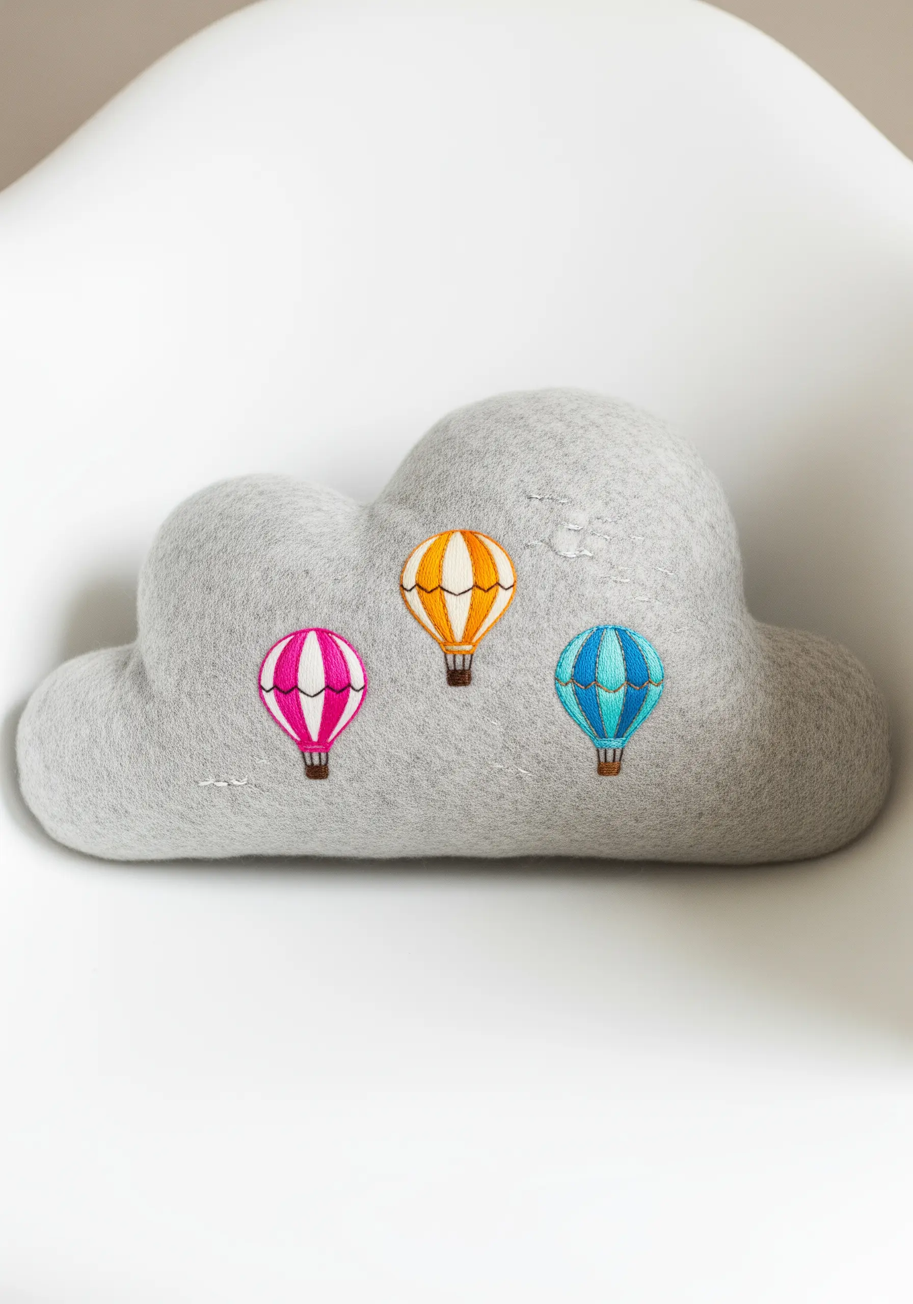 Three small, colorful hot air balloons embroidered onto a grey felt cloud-shaped pillow.