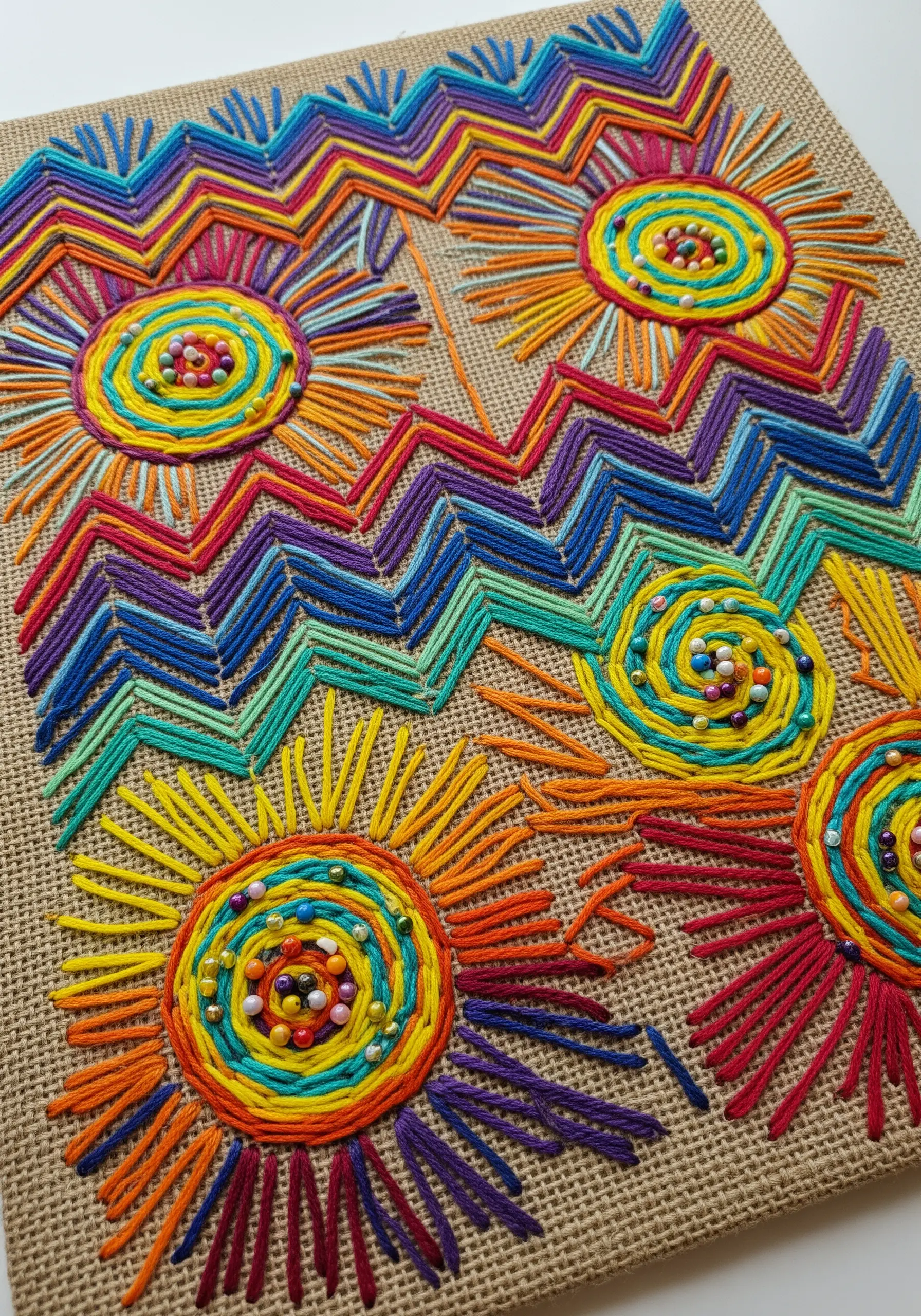 Colorful yarn embroidery on burlap with multicolored seed beads in the center of spiral suns.