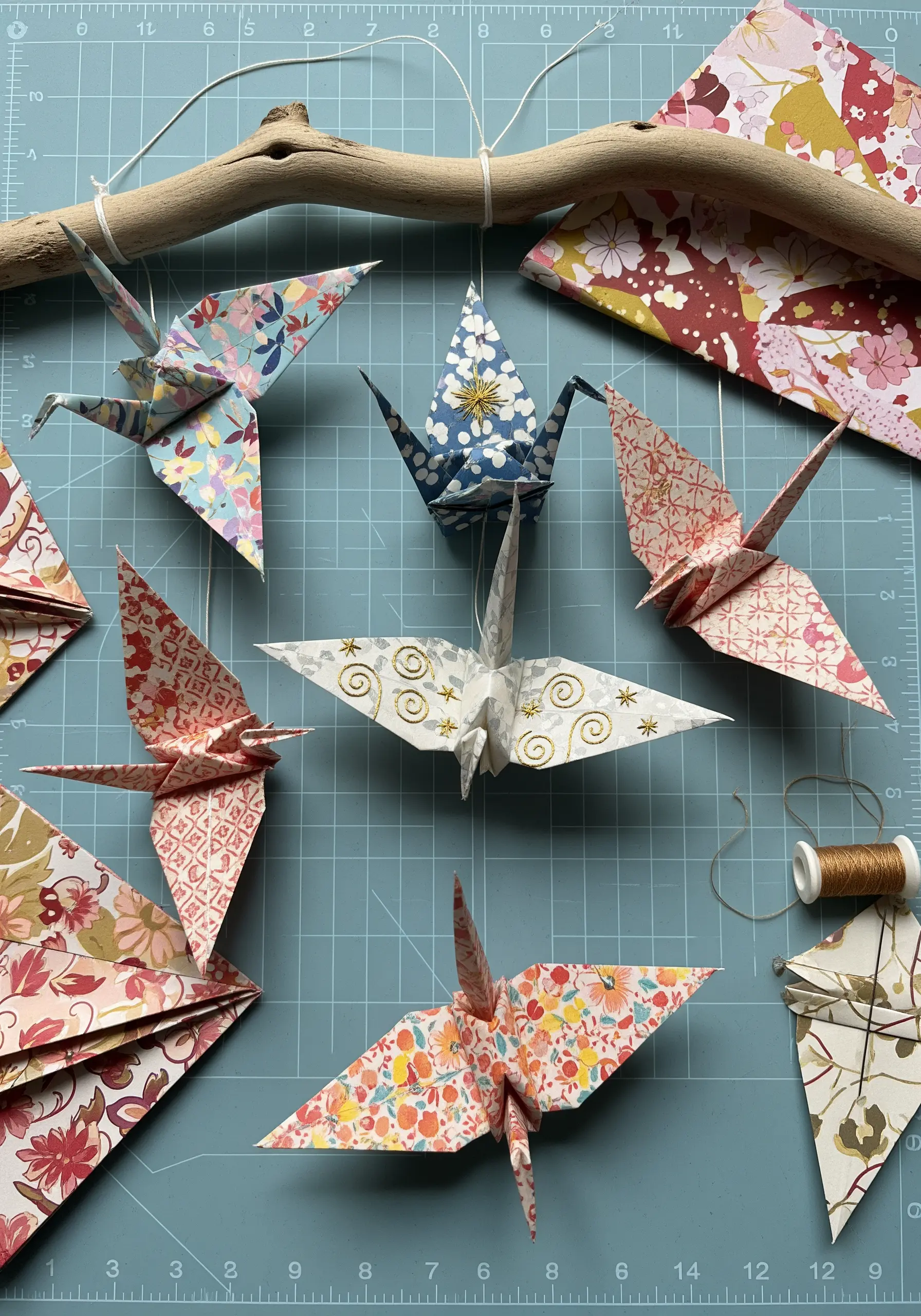 Origami paper cranes with subtle gold thread details, hanging from a branch.