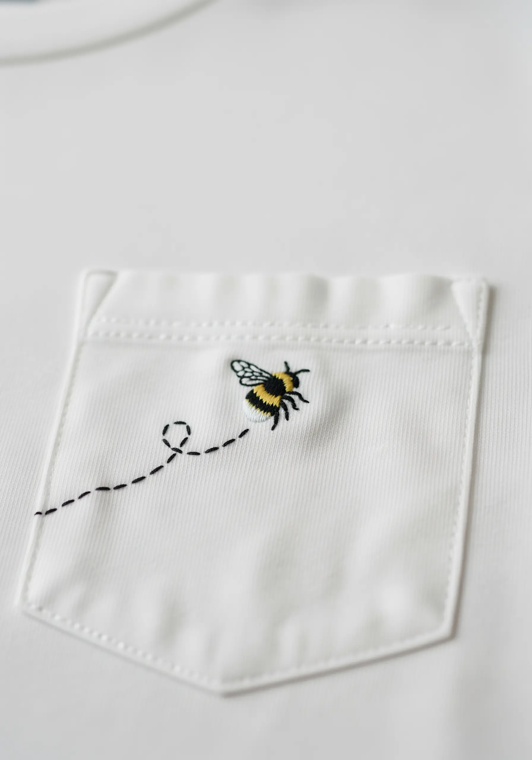 A small embroidered bee with a dashed-line flight path on a white T-shirt pocket.