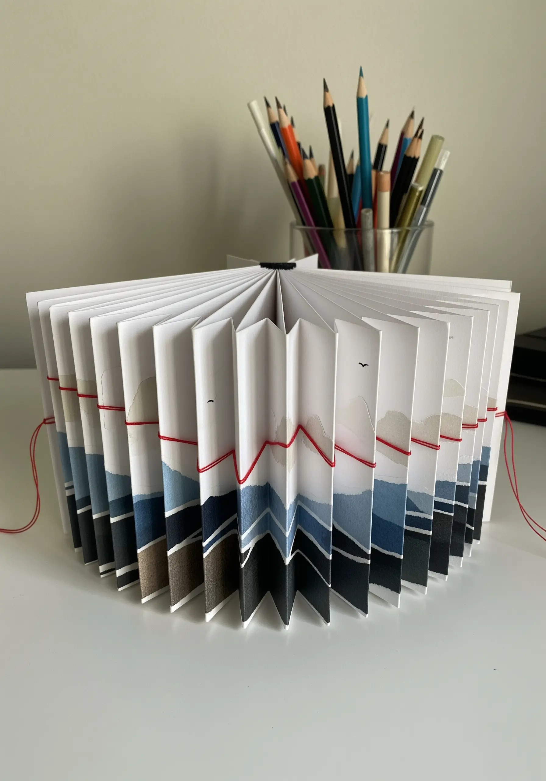 An accordion-folded paper book with a painted landscape, bound together with a red thread.