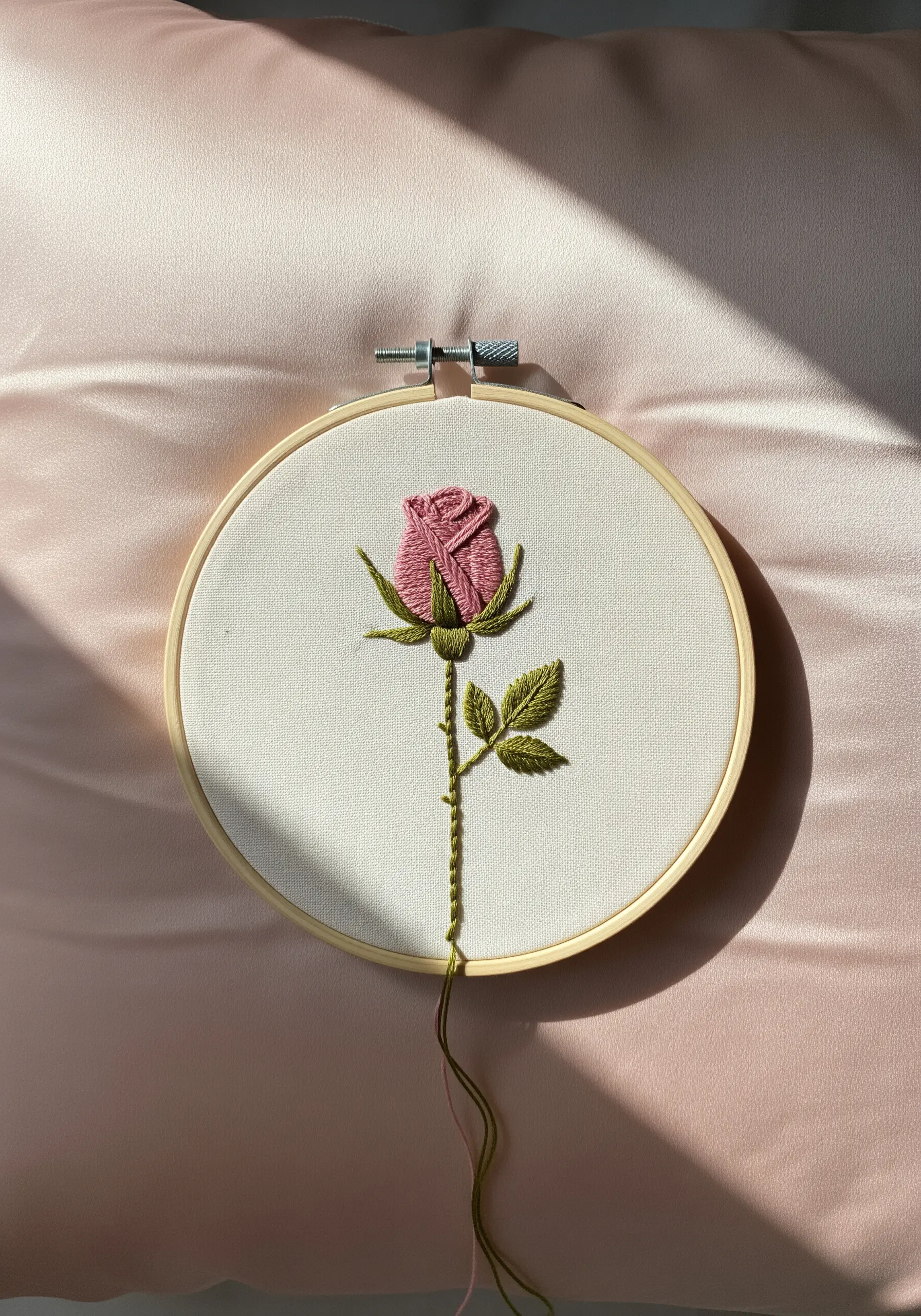 Thread-painted pink rose with green leaves on off-white fabric, displayed on a satin pillow.