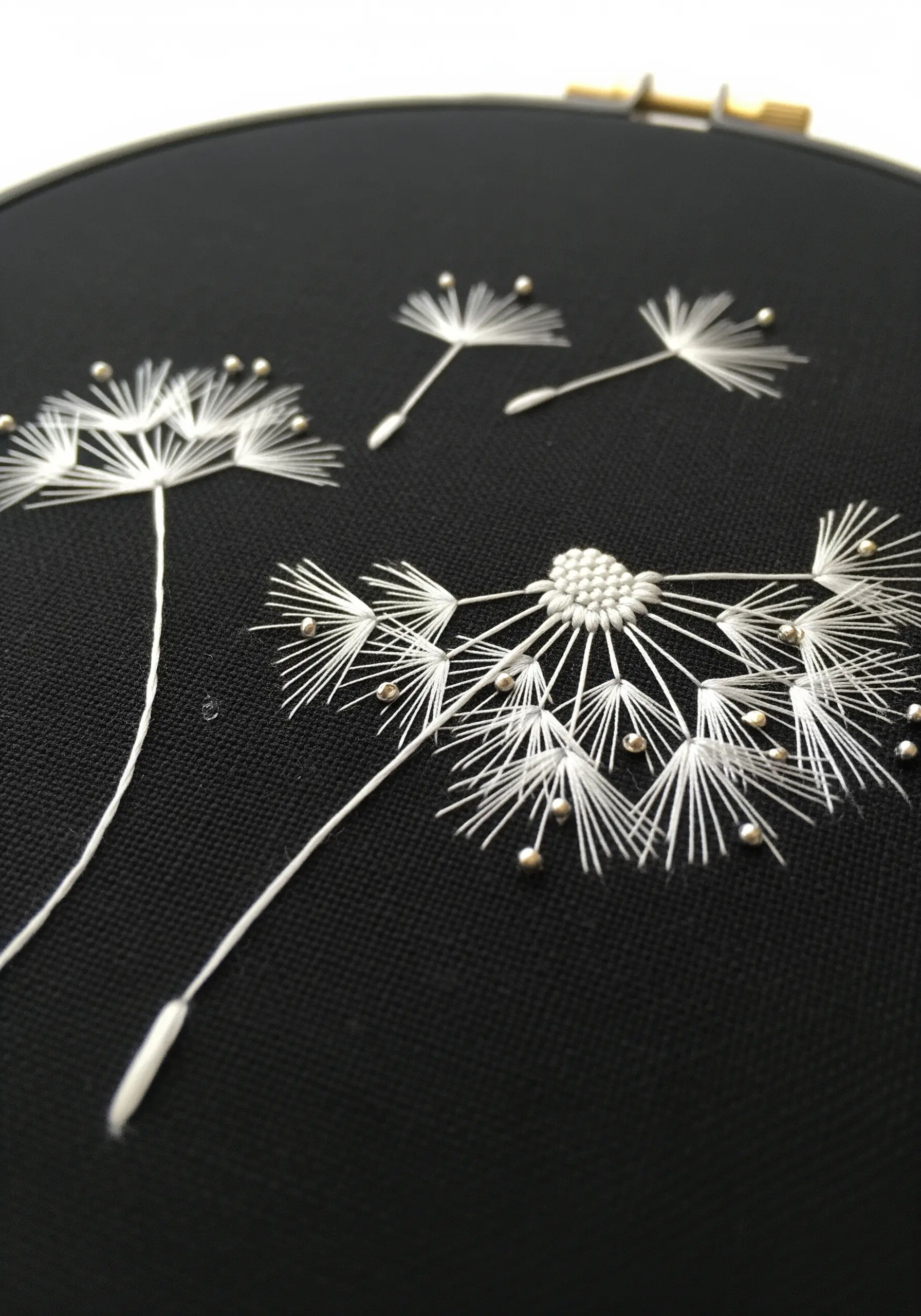 Delicate dandelion seed heads embroidered in white thread with silver beads on black fabric.