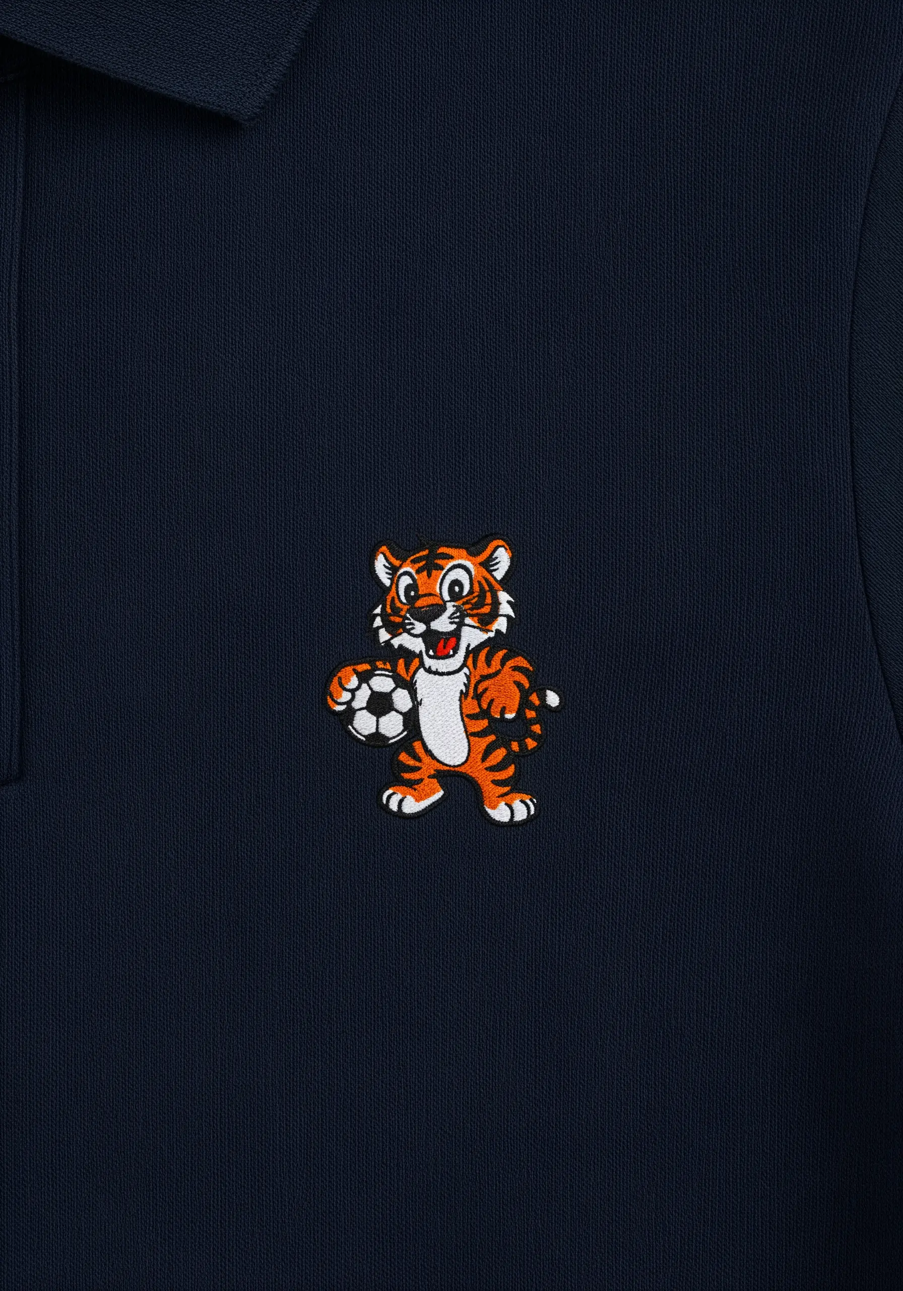 A small, detailed cartoon tiger holding a soccer ball, embroidered on a dark blue polo shirt