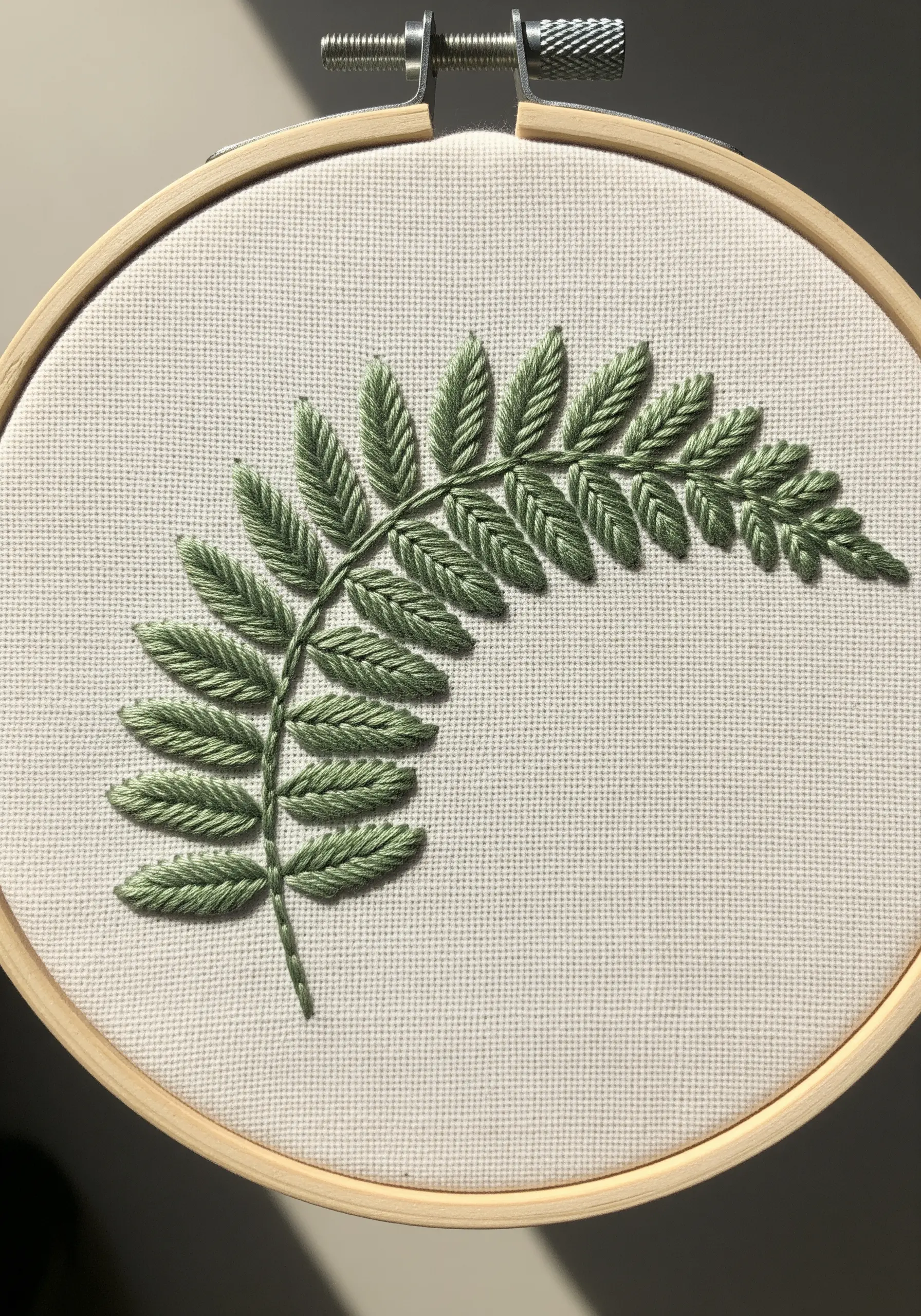 A single fern frond embroidered in sage green using fishbone stitch on cream fabric.