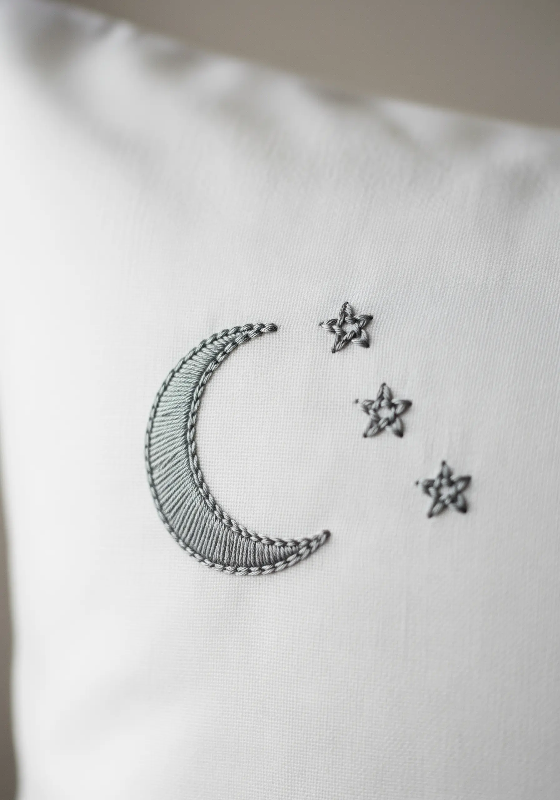 A crescent moon and three stars embroidered in grey thread on a white pillowcase.