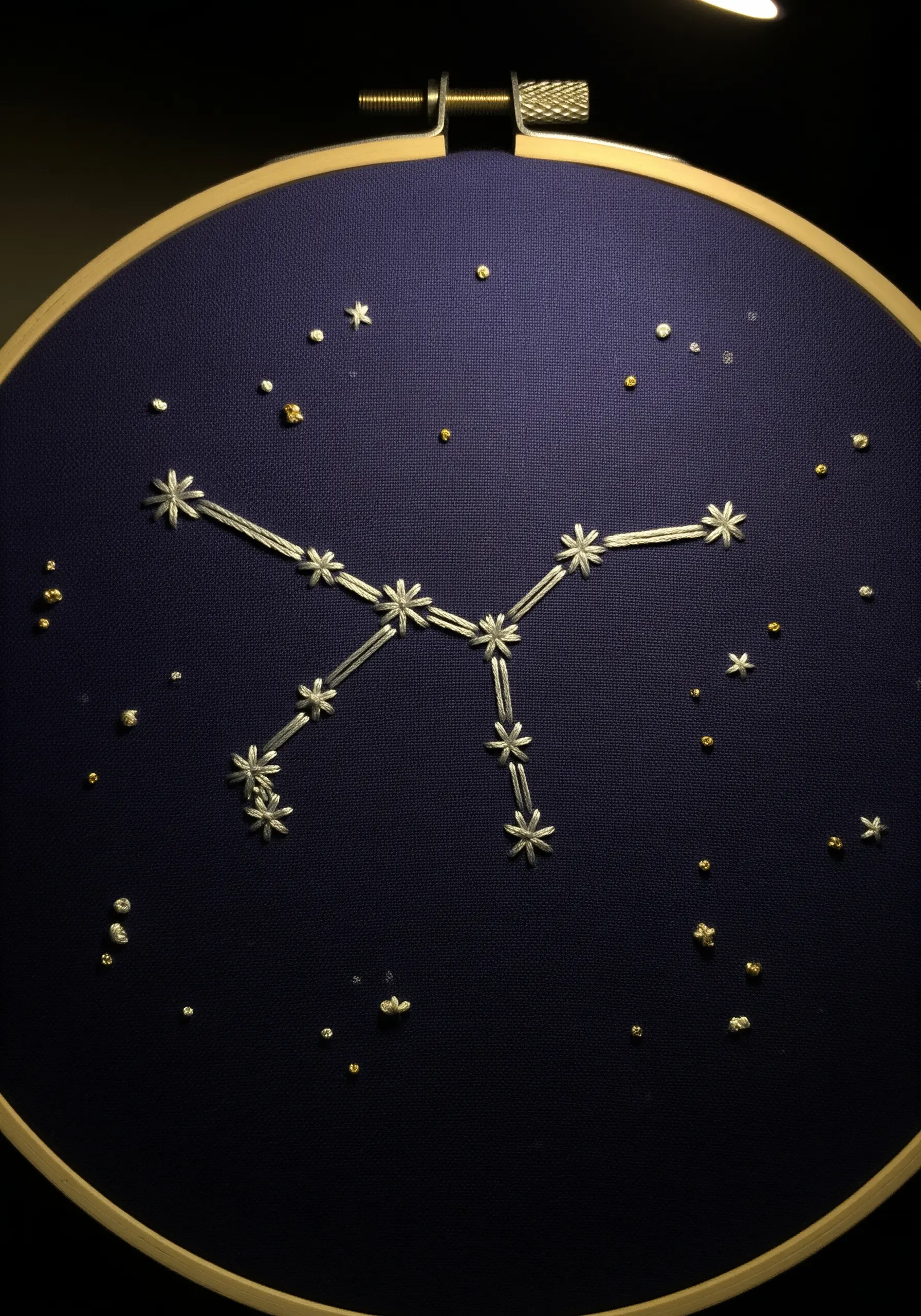 A simple embroidered constellation with star stitches and French knots on dark navy fabric in a hoop.