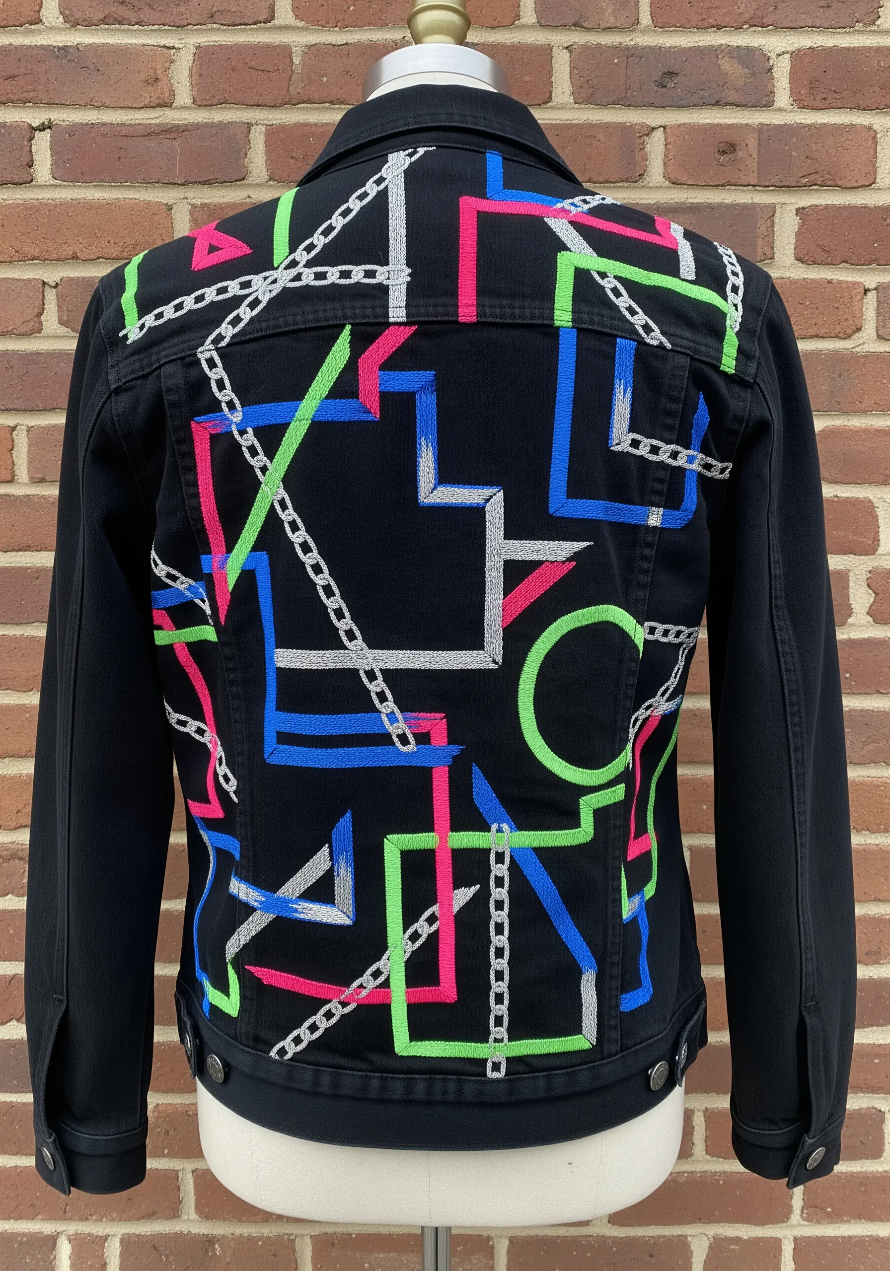 A black denim jacket with abstract geometric embroidery in neon pink, blue, and green.