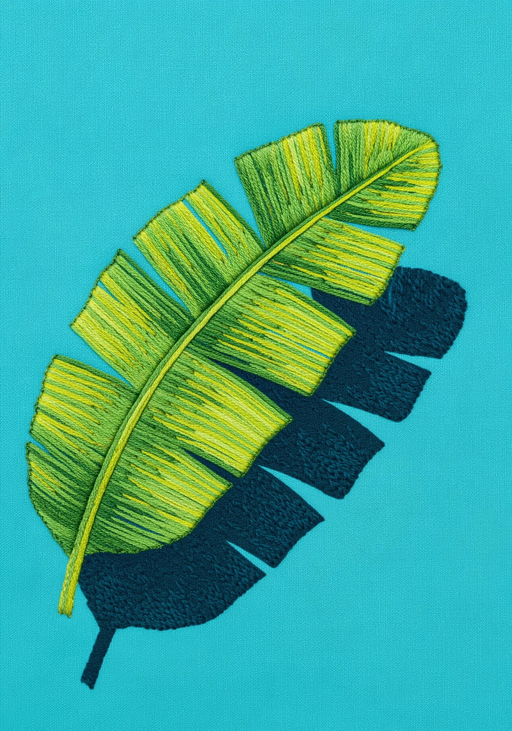 A green and yellow banana leaf embroidered on bright turquoise fabric with a navy blue shadow.