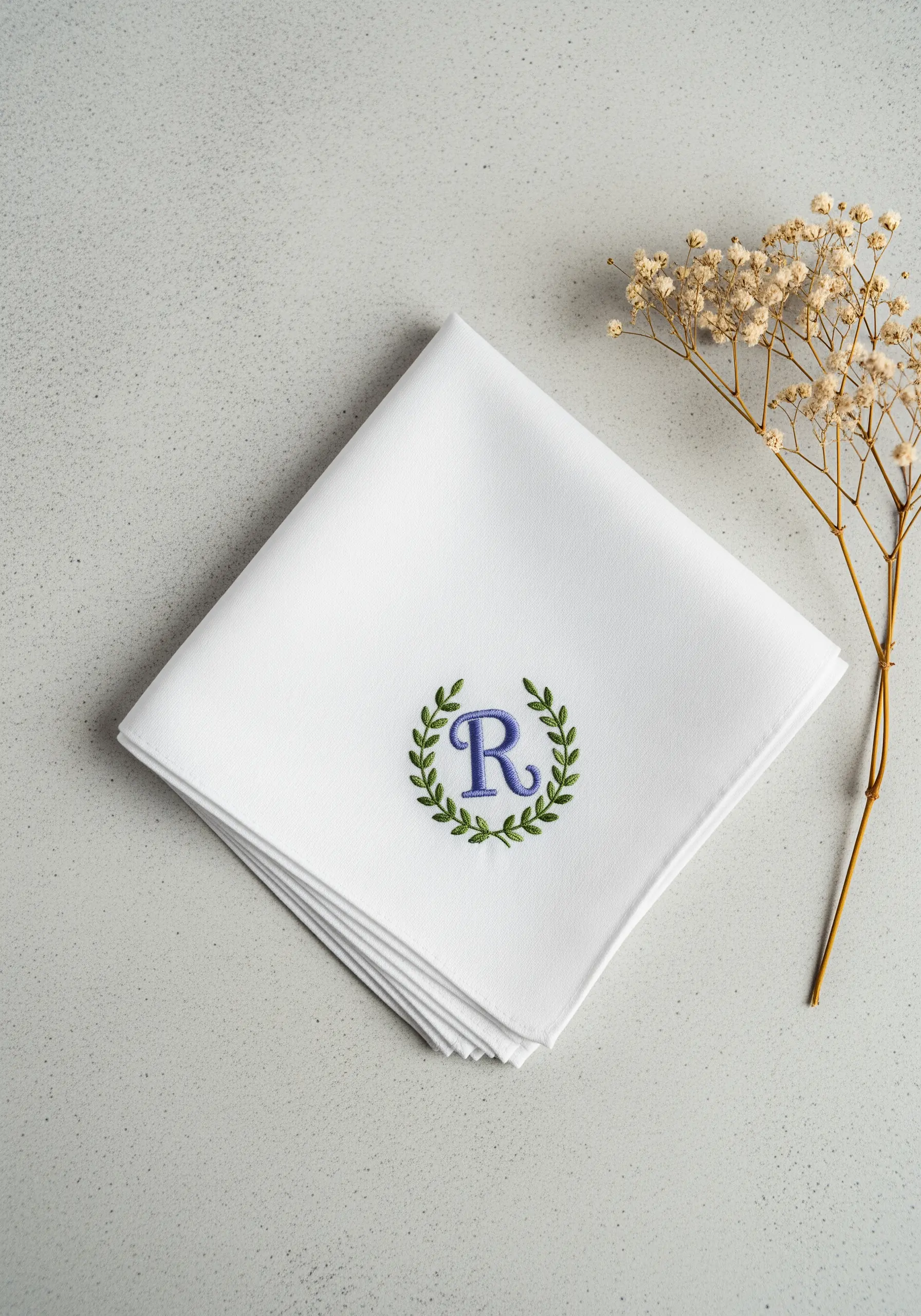A crisp white handkerchief with an embroidered blue letter 'R' inside a green laurel wreath