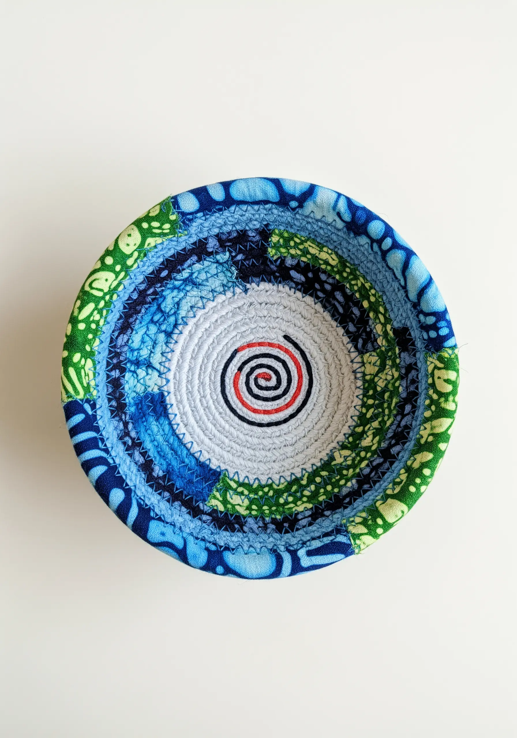 A colorful coiled rope bowl made with blue and green patterned fabrics, with a red spiral inside.
