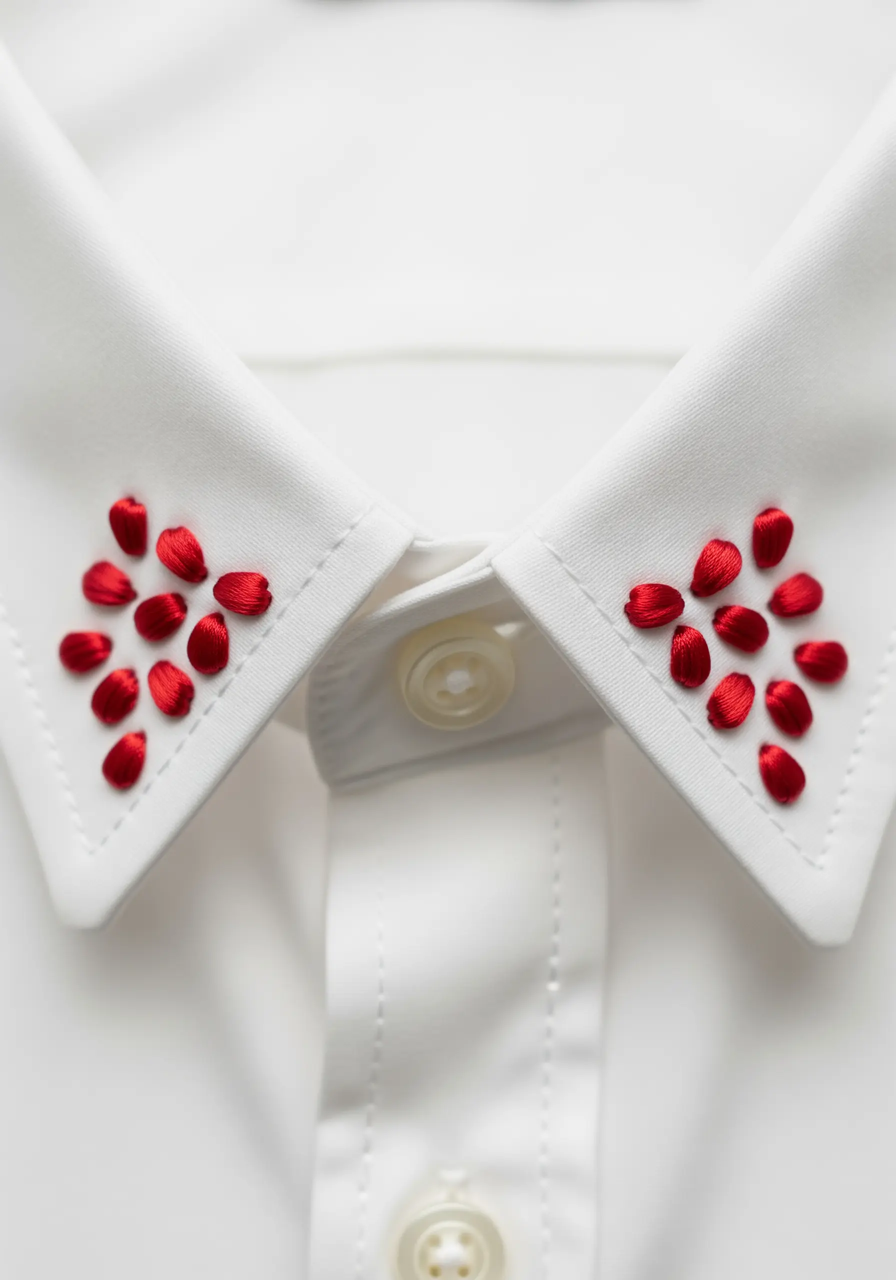 Scattered red satin stitches resembling pomegranate seeds on the tips of a white shirt collar.