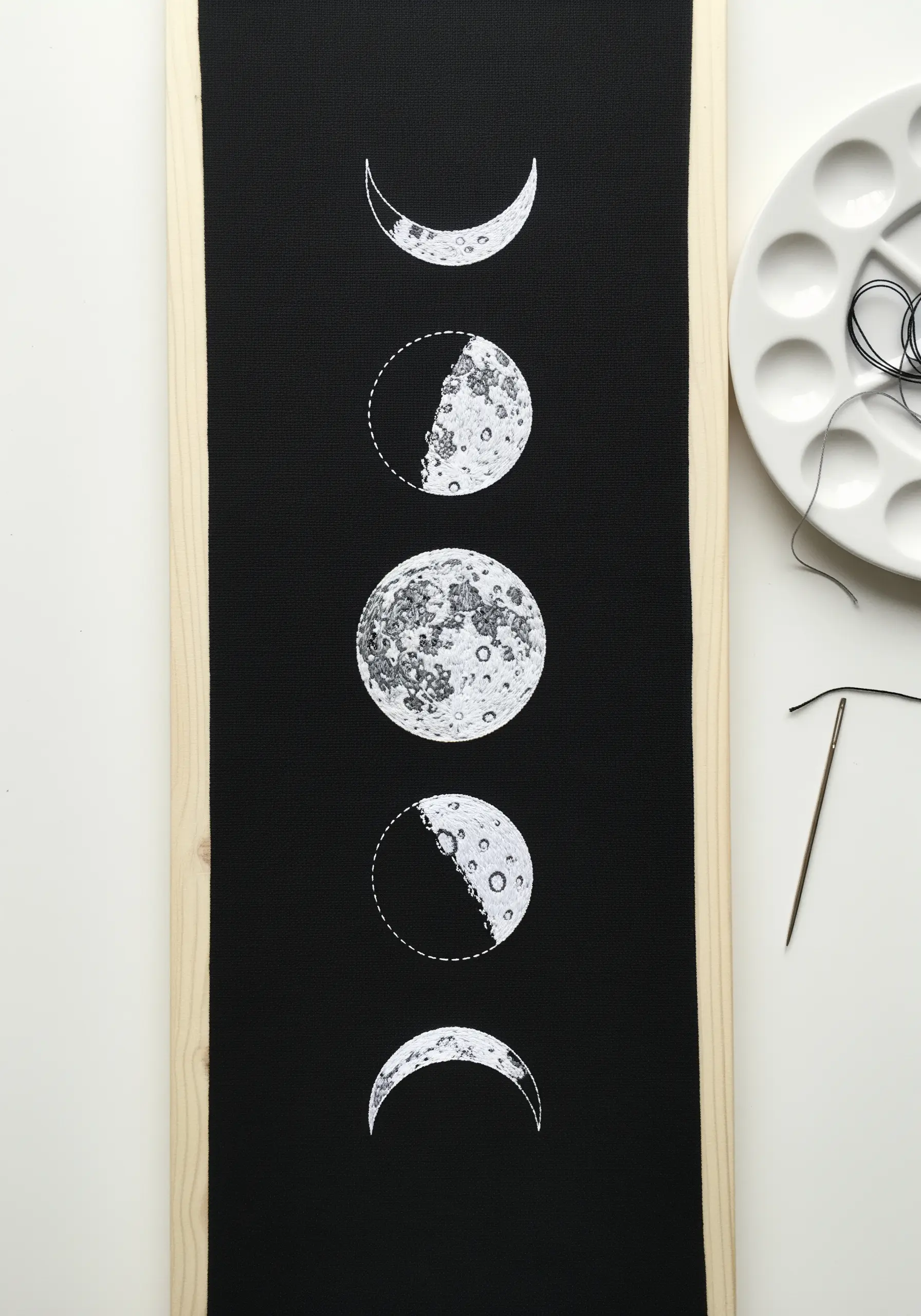 A vertical embroidery of the phases of the moon on black fabric, combining paint and white thread.