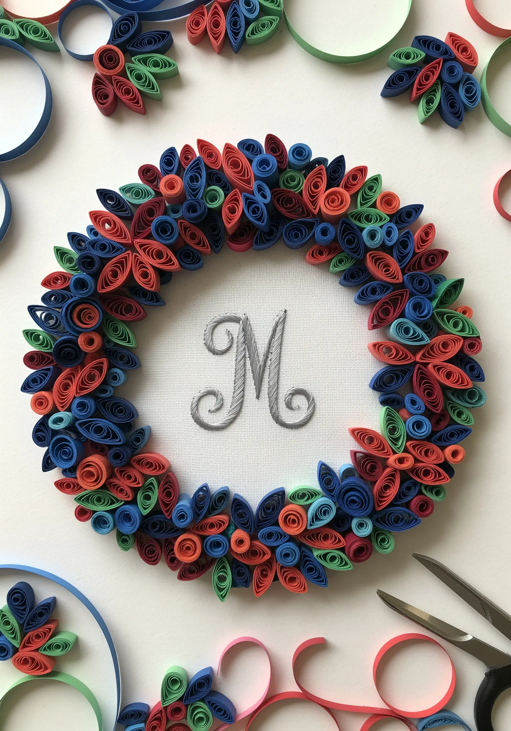 A gray embroidered monogram 'M' centered within a vibrant paper quilling wreath.