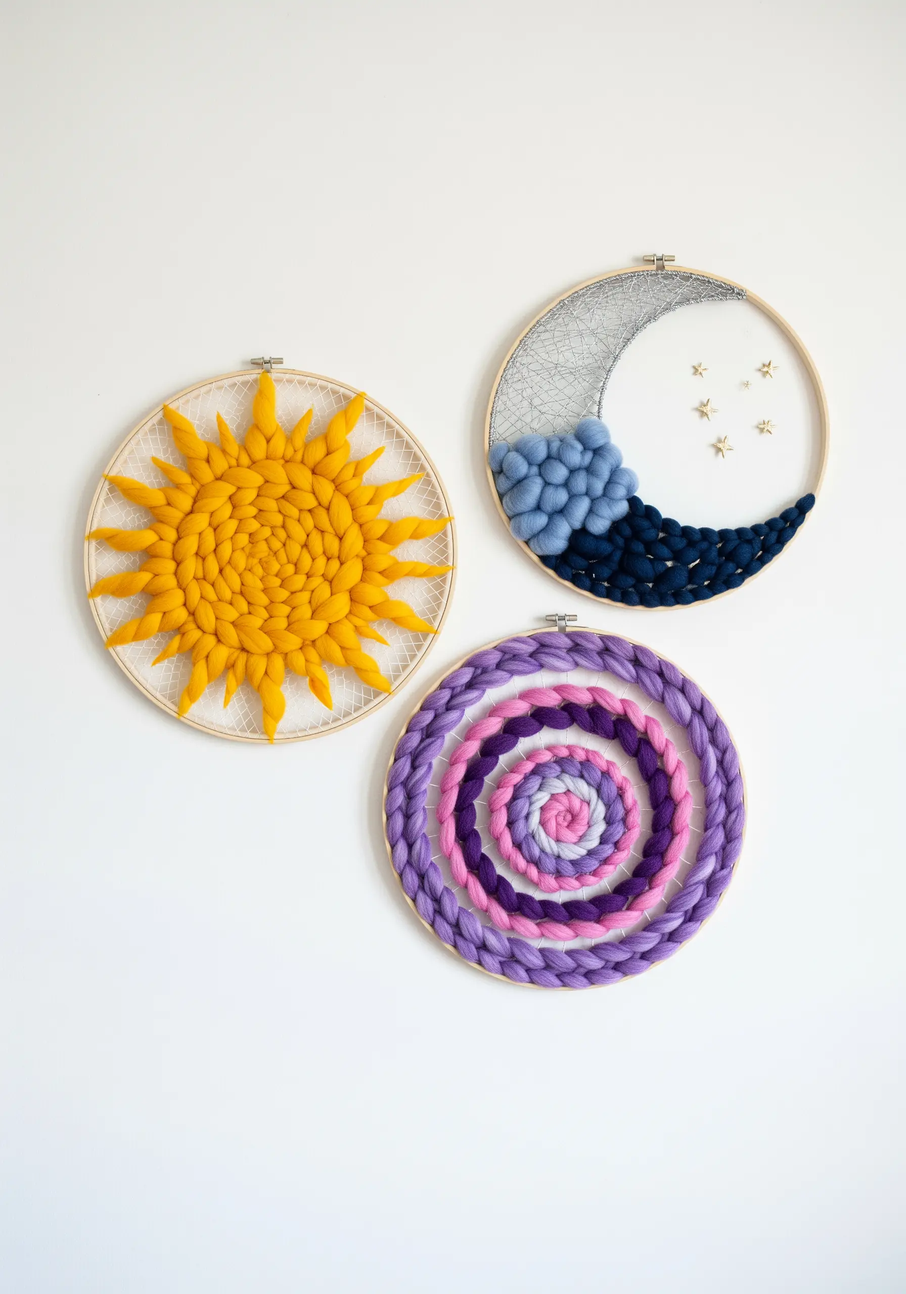 Three fiber art hoops: a woven sun, a felted moon and clouds, and a braided purple spiral.