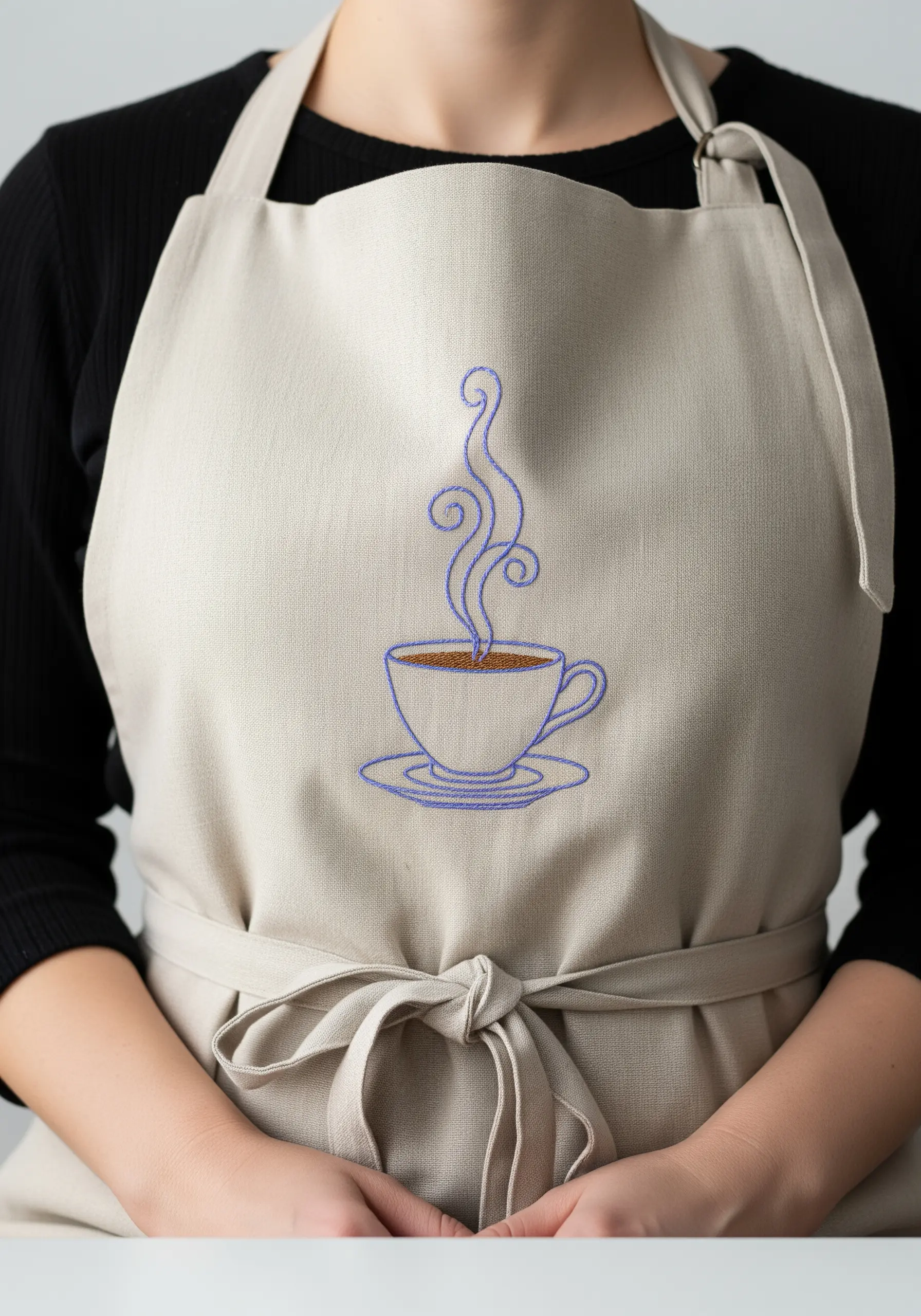 A line art coffee cup embroidered in blue thread on a neutral-colored linen apron.