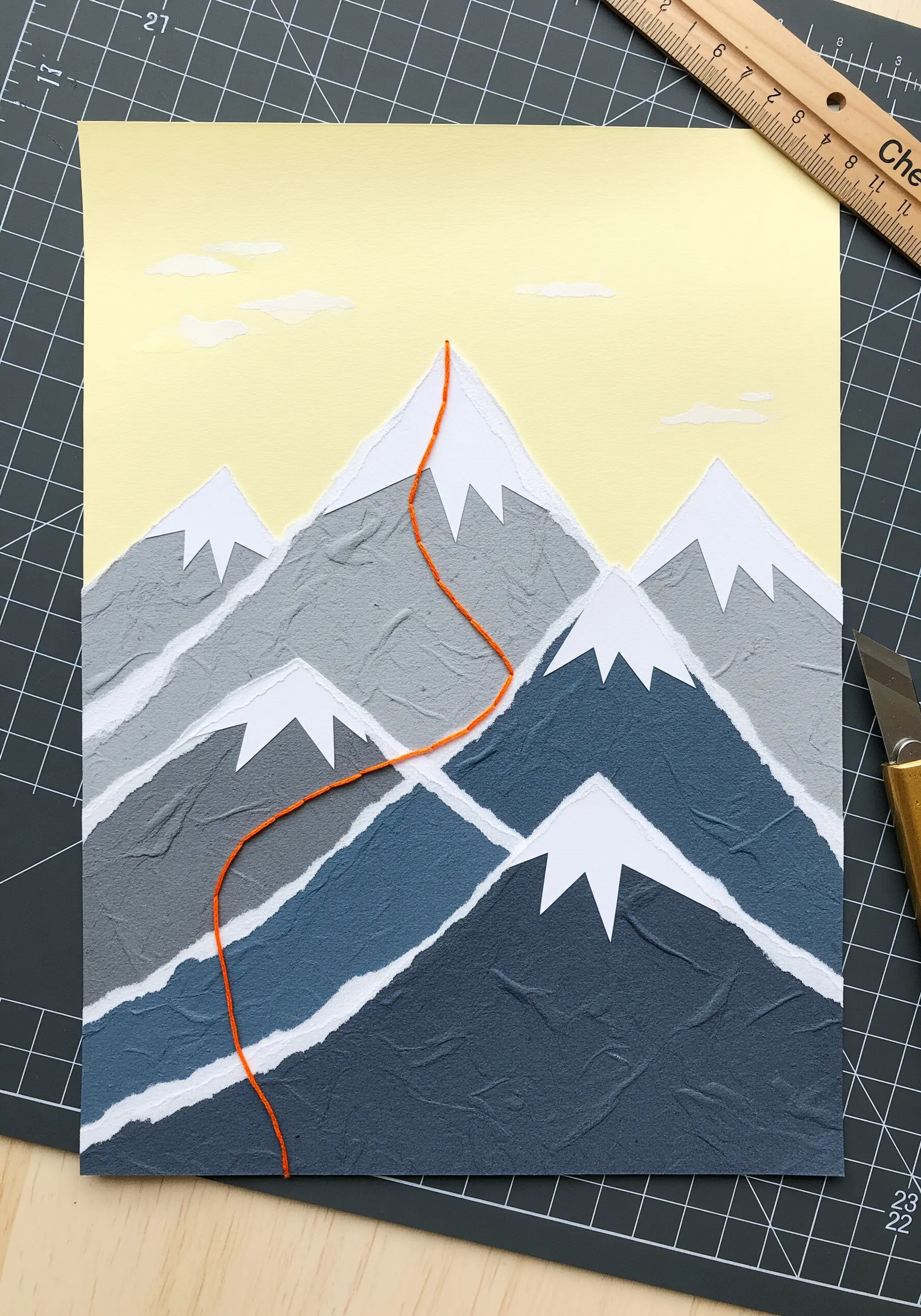 Torn paper collage of a mountain range with a bright orange couched yarn path leading up.