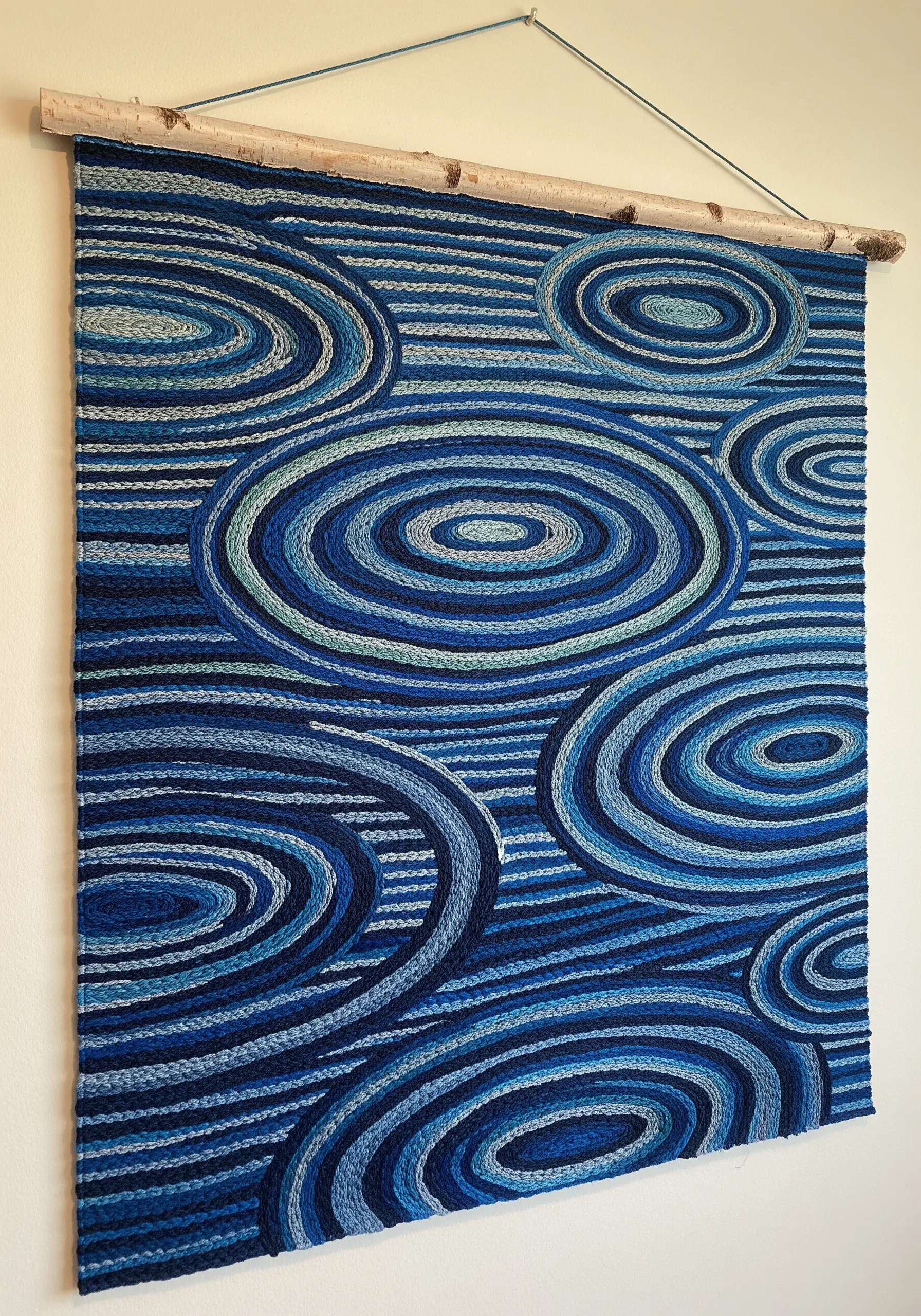 Large wall hanging with couched blue yarn creating concentric water ripple patterns.