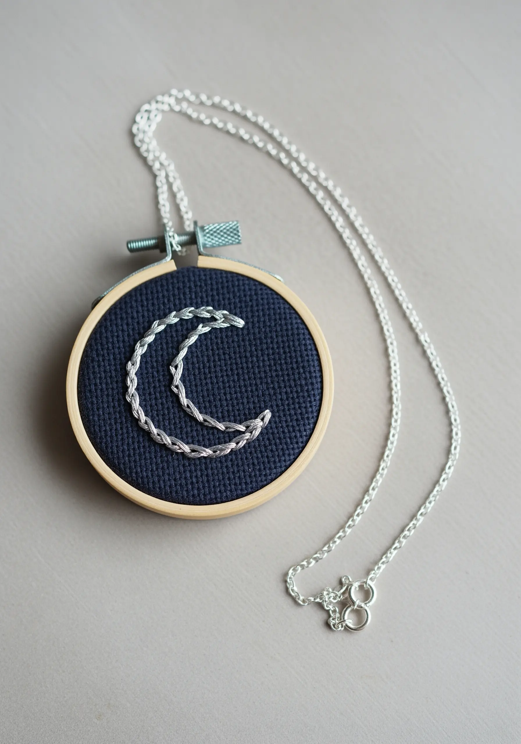 A crescent moon embroidered with silver chain stitch on dark blue fabric in a mini hoop necklace.