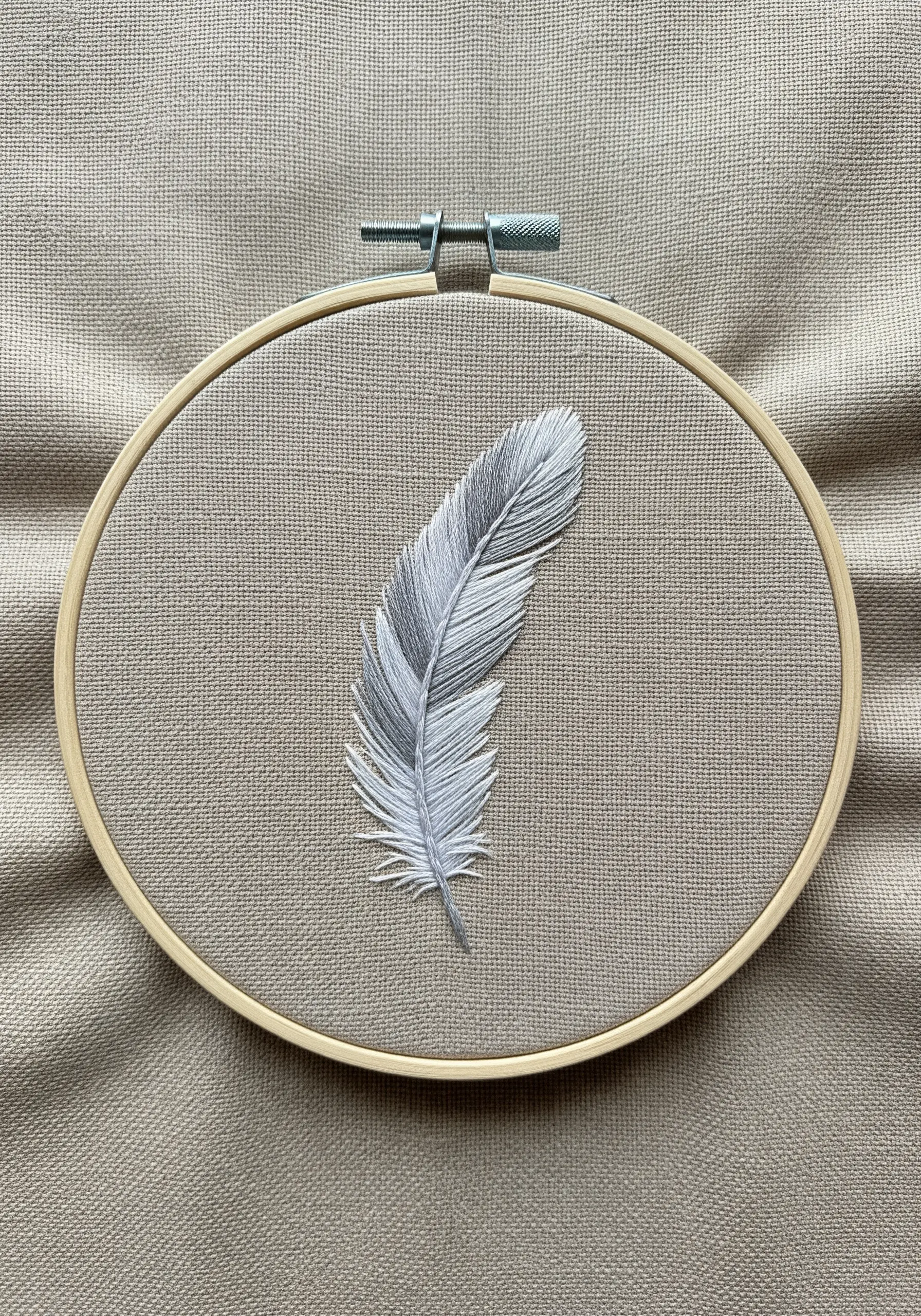 A realistic white and gray feather embroidered with angled stitches on neutral fabric.
