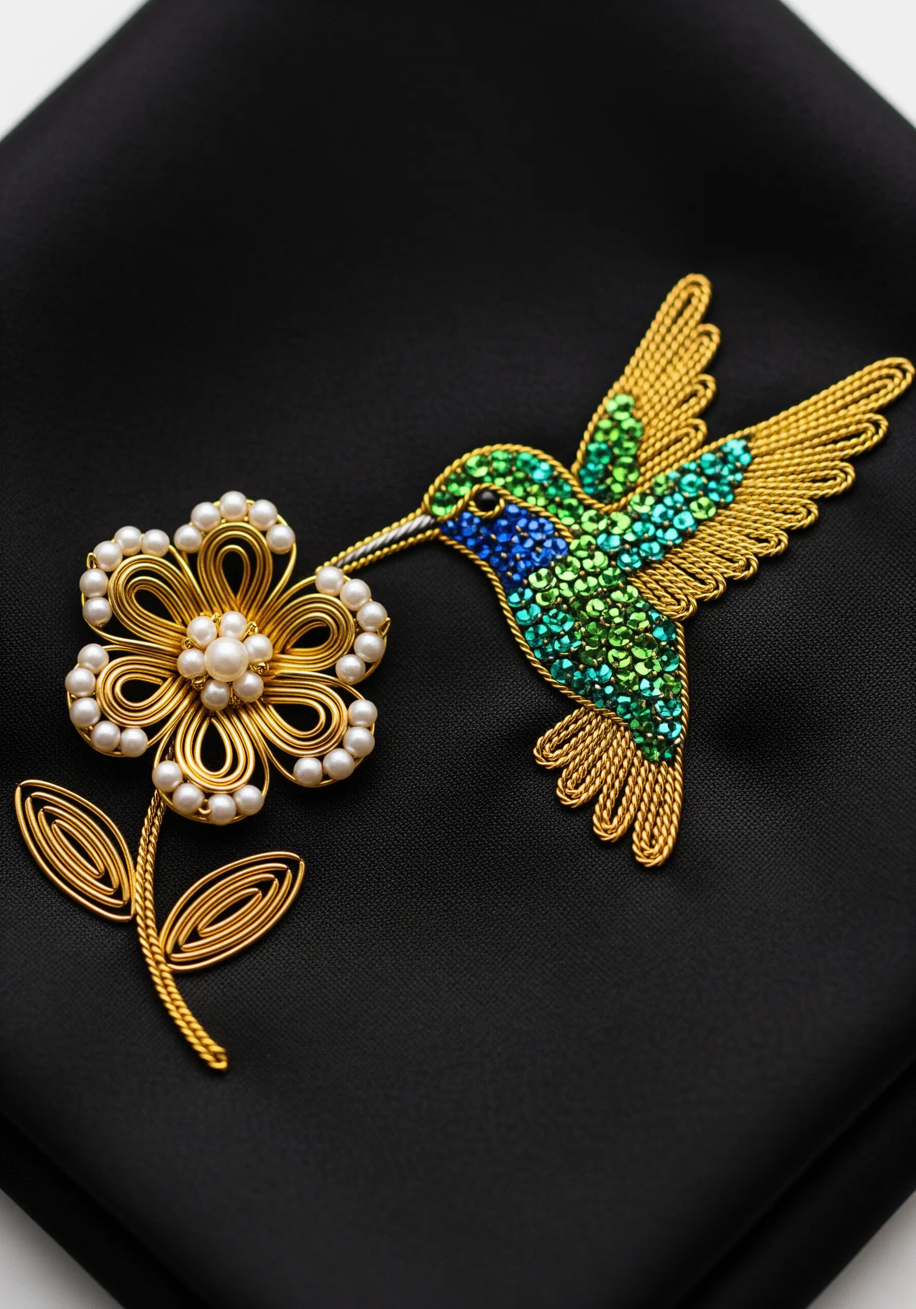 A hummingbird and flower made of gold wire, rhinestones, and pearls on black fabric.
