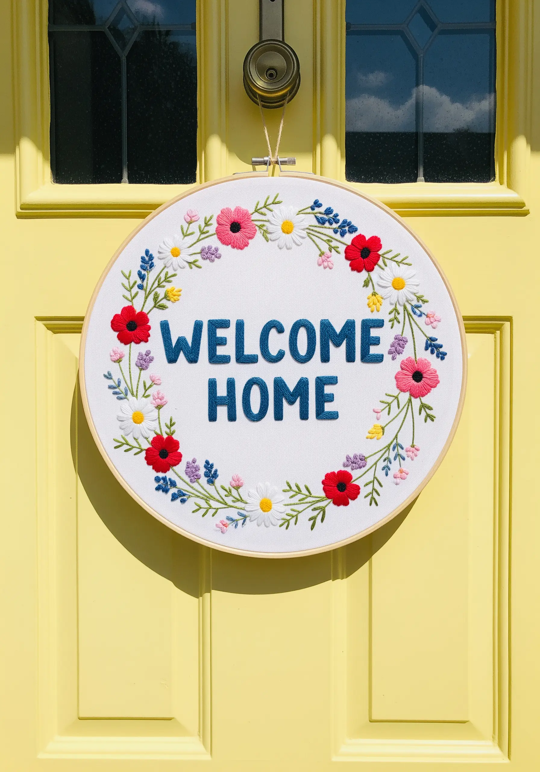 A 'Welcome Home' embroidery hoop wreath with colorful flowers hanging on a yellow door.