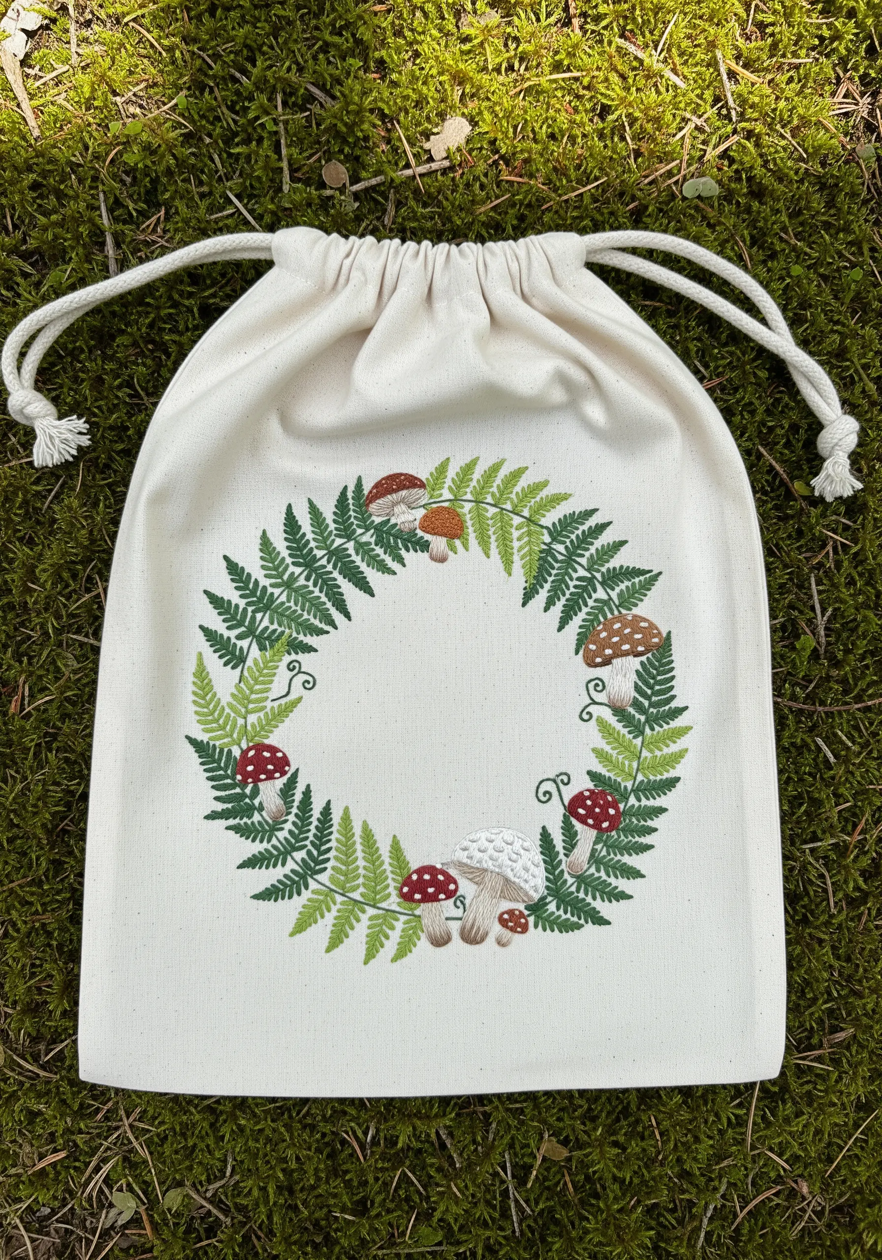 An embroidered wreath of ferns and mushrooms on the front of a canvas drawstring bag.