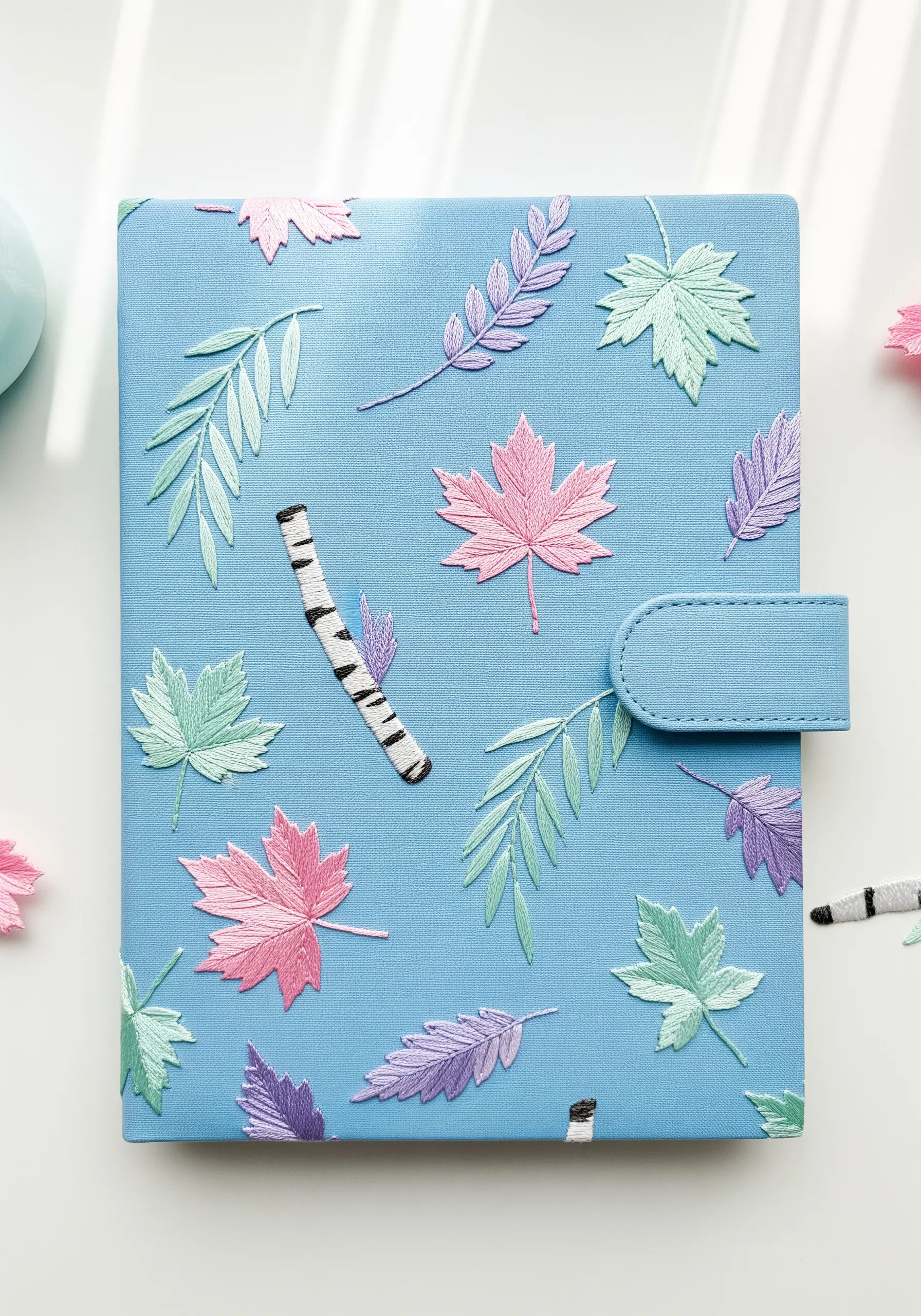 A scattered pattern of pastel-colored embroidered leaves and birch logs on a blue journal.