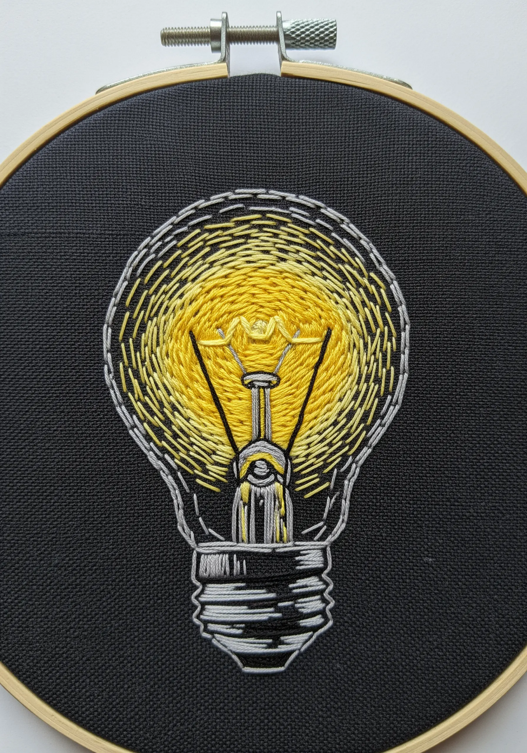 An embroidered lightbulb on black fabric with a radiating yellow and white glow effect.