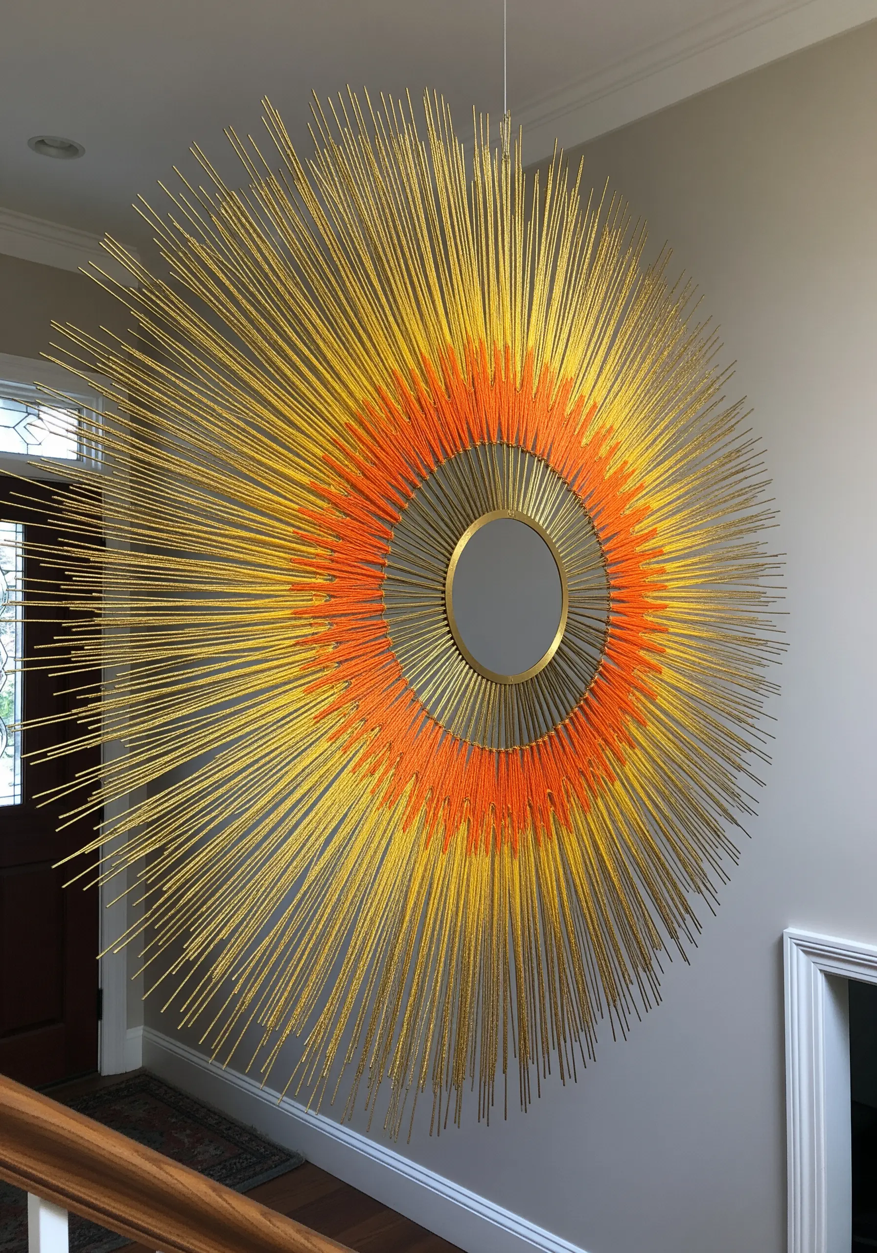 A large sunburst wall sculpture with a central mirror and radiating spokes wrapped in yellow and orange yarn.