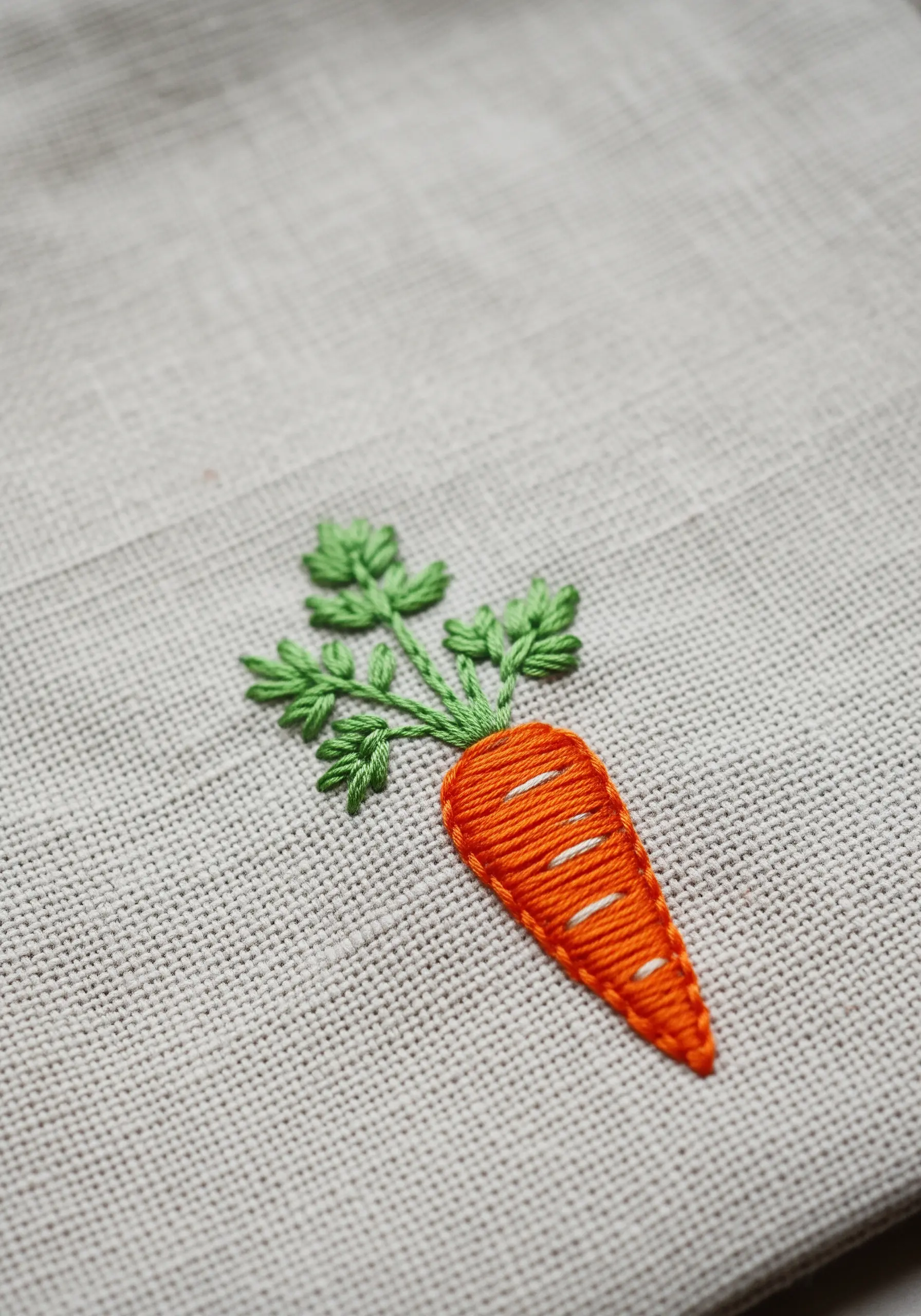 A small embroidered carrot with a raised satin stitch body on linen fabric.