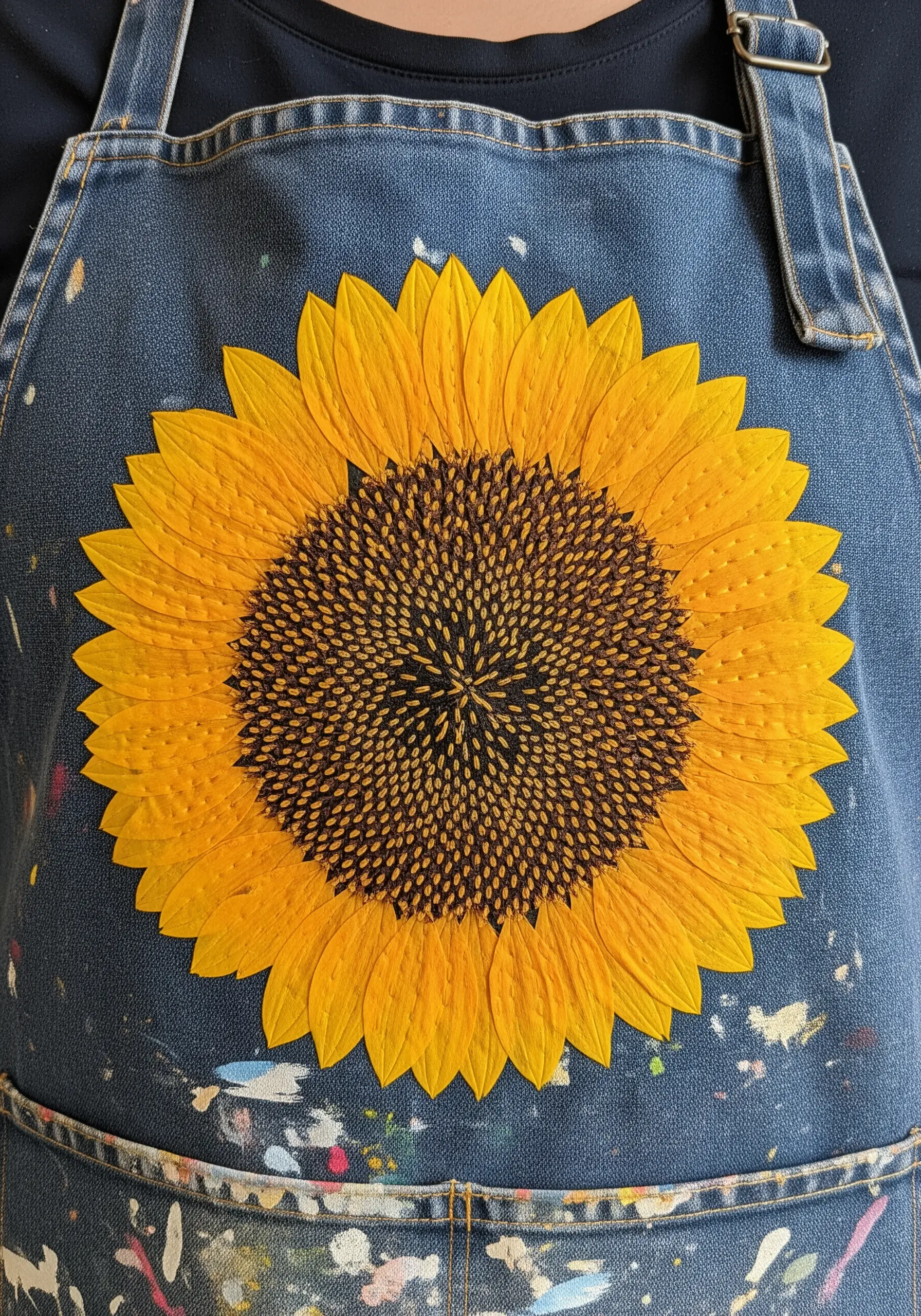 A large sunflower made from fabric appliqué petals and a dense knot-stitched center on a denim apron.