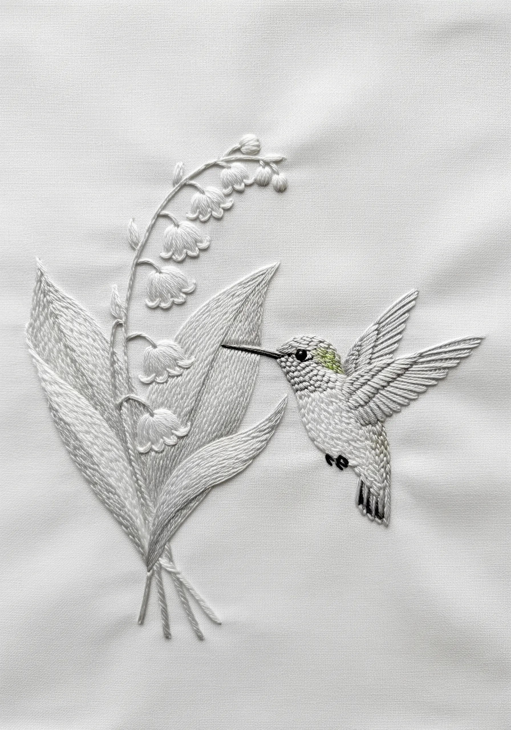 A white hummingbird and lily of the valley embroidered in white thread on white fabric.