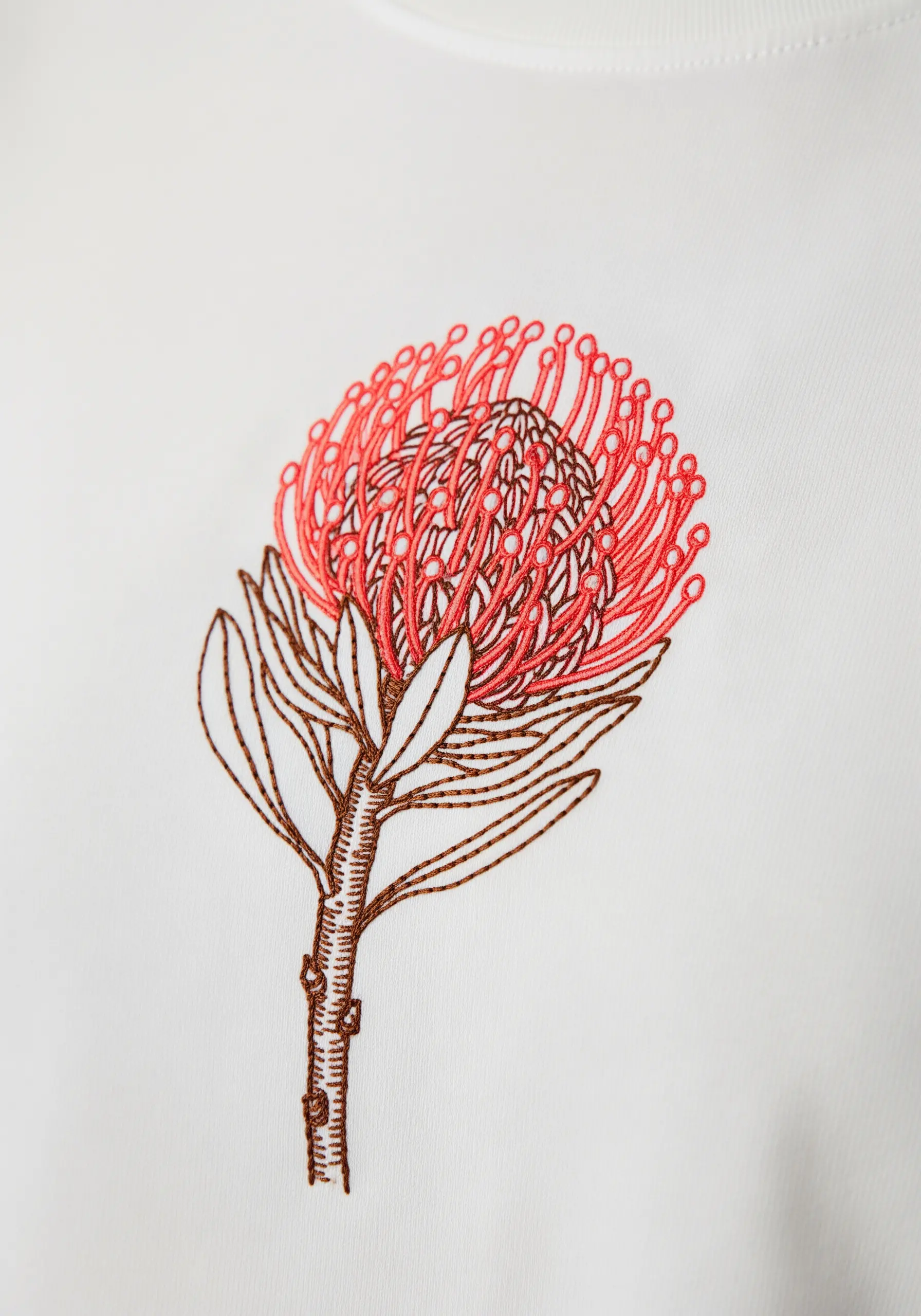 A protea flower embroidered with red and brown line art on a white sweatshirt.